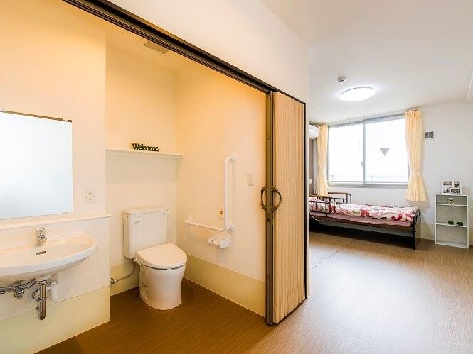 トイレ付きの居室には寝具付きのベッドが置かれ、大きな窓もある。トイレにはアコーディオンカーテンが付いている。