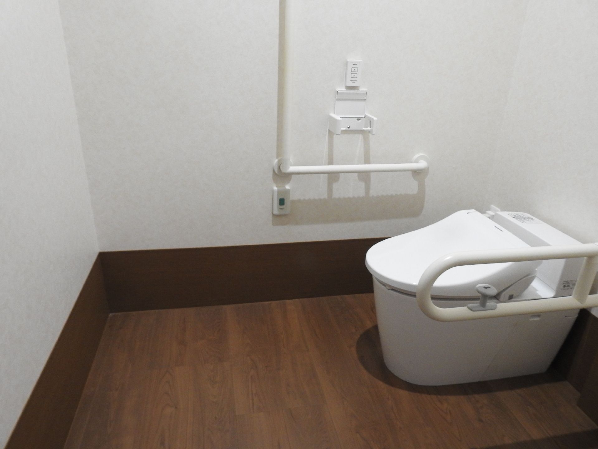白を基調としたトイレ。フローリングの床には何もなく、シンプルなトイレの両サイドに手すりが付き、壁にはトイレットペーパーホルダーもある。