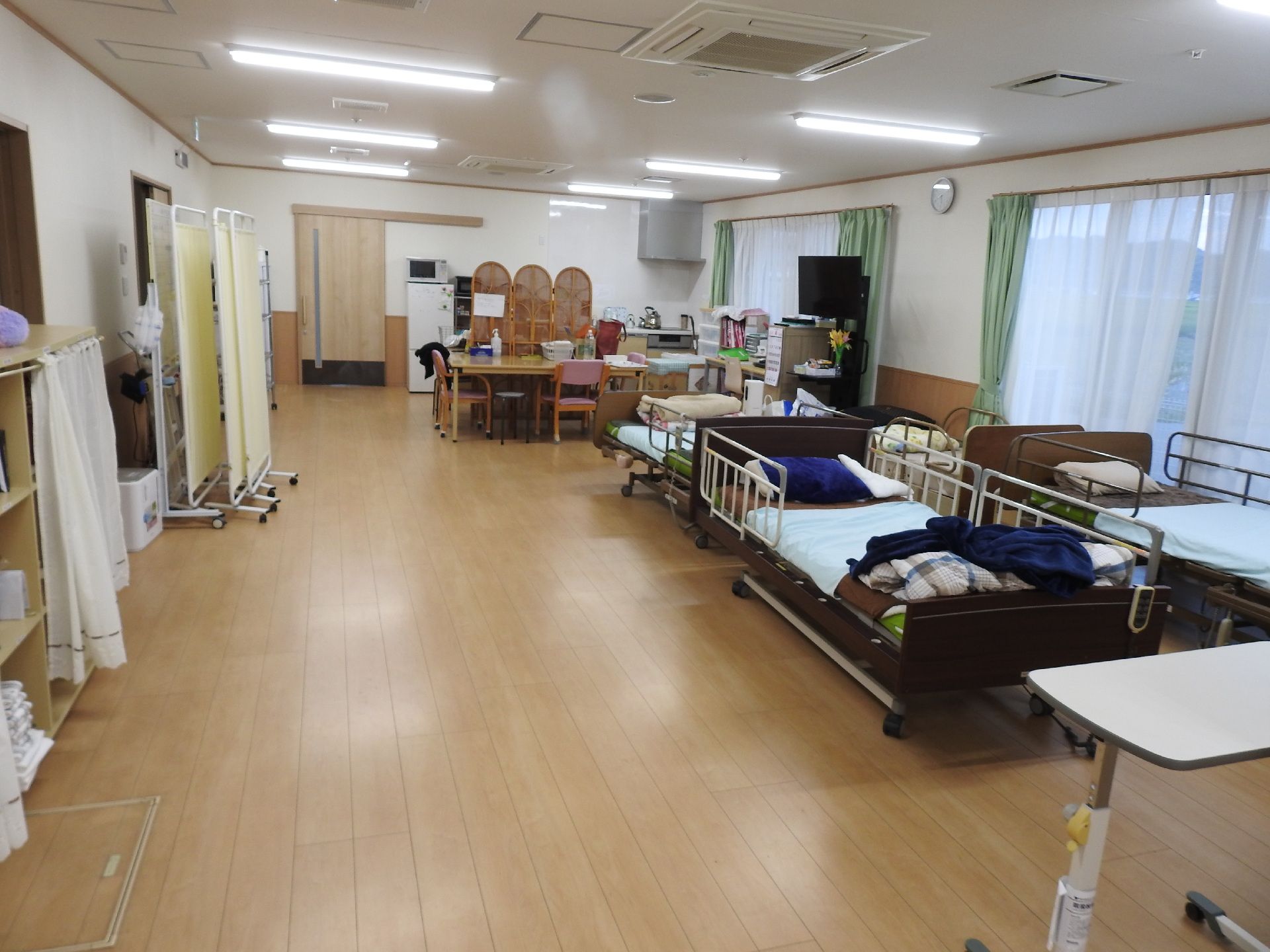 介護ベッドが複数窓際に置かれた共有スペース。黄色いカーテン式のパーテーションが左側にあり、奥にはテーブルセットやテレビもある。