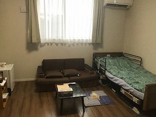 レースカーテンとカーテンが付いた大きな窓があるフローリングの部屋にはソファやベッド、テーブルなどが置かれている。
