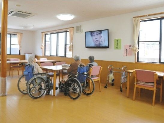 大きなテレビが壁に設置された食堂。テーブルを囲んでいる車いすの高齢女性たちが見ている。