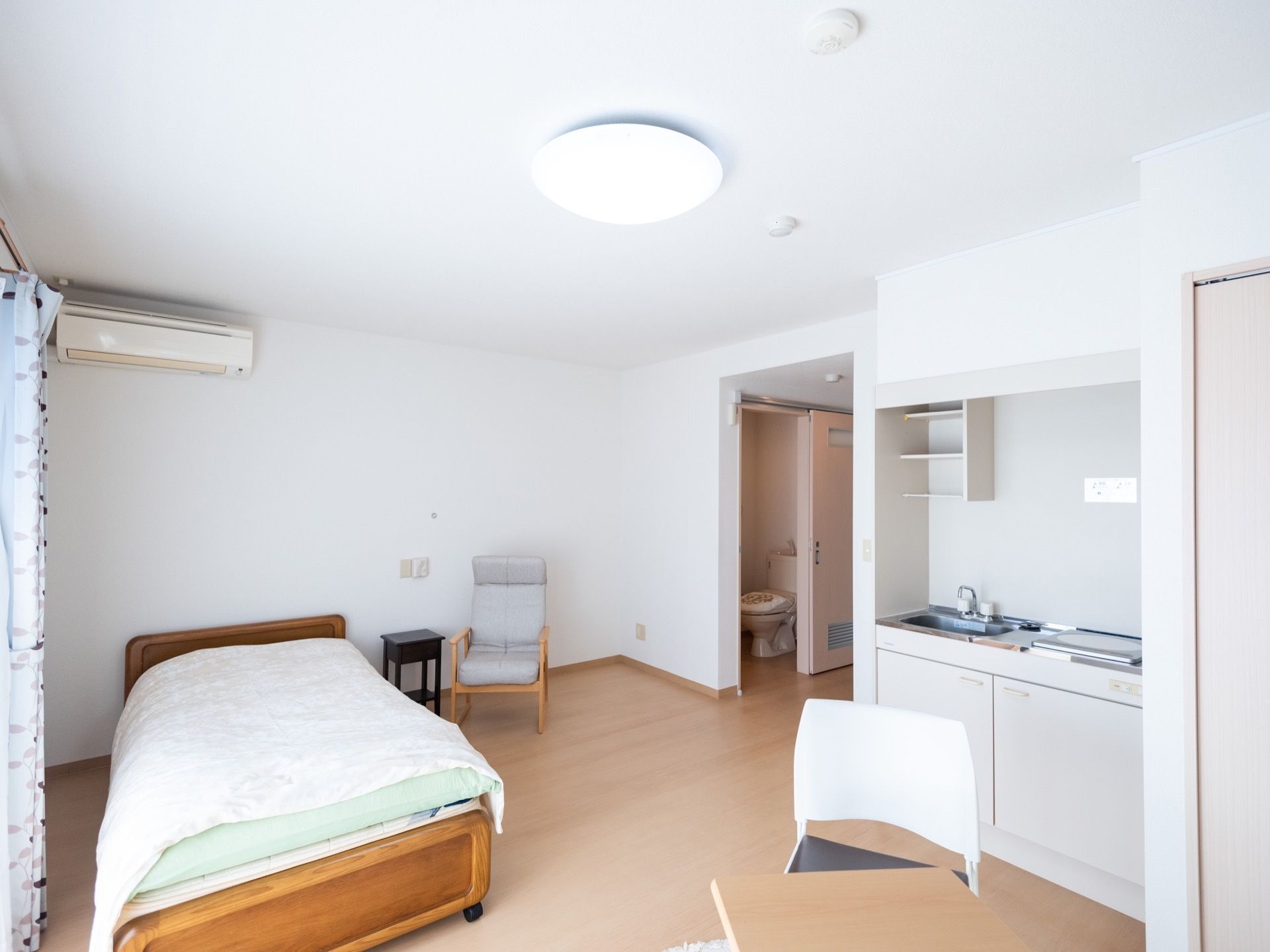 清潔感のある白で統一された美しい居室である。トイレやキッチン、ベッド、エアコンといった生活に最低限な設備や家電、家具が取り揃えられている。