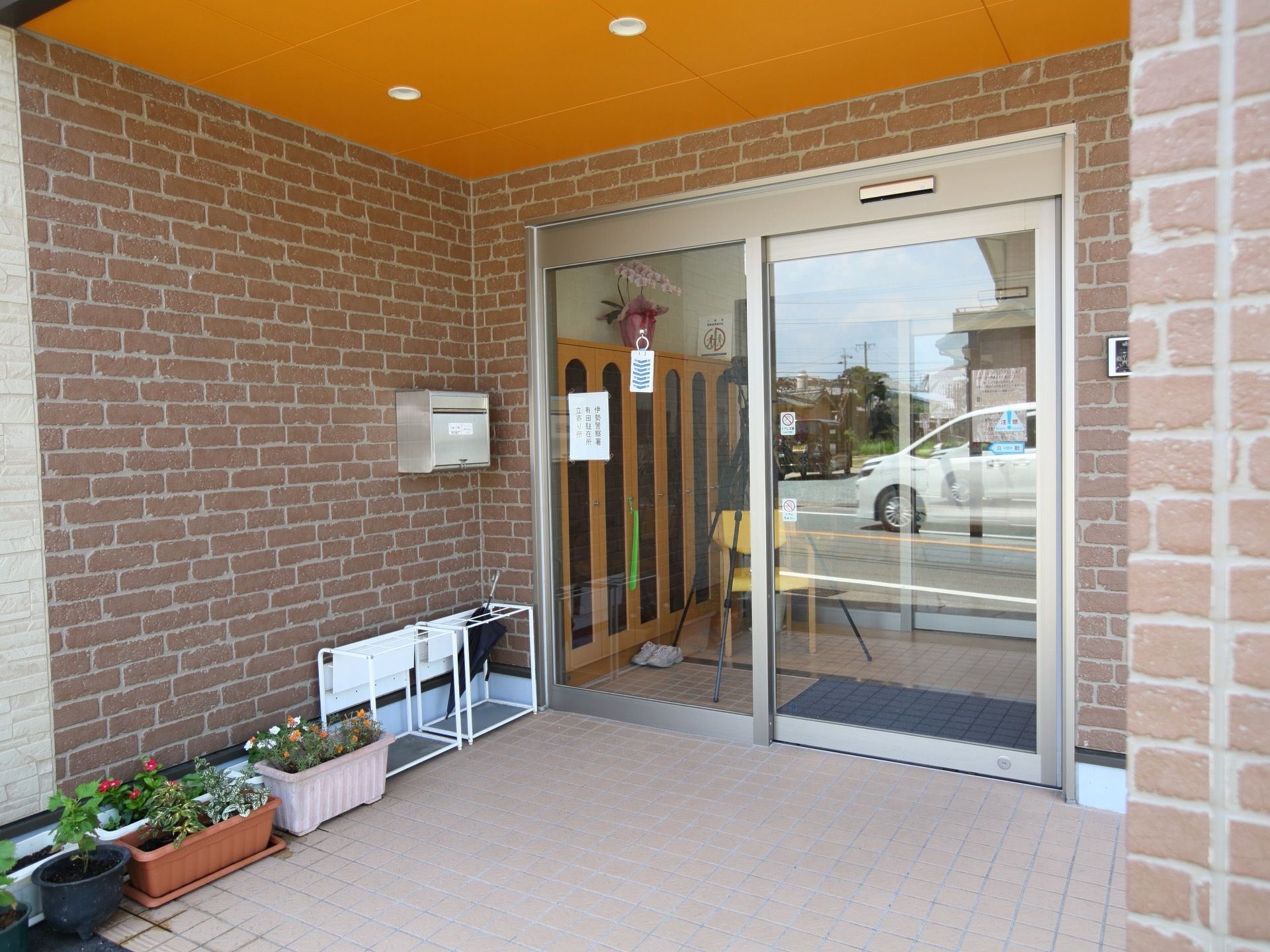 大きなひさしのある玄関。玄関の扉は自動ドアとなっていて、玄関の周囲には傘立てや花の植えられた植木鉢などが置かれている。