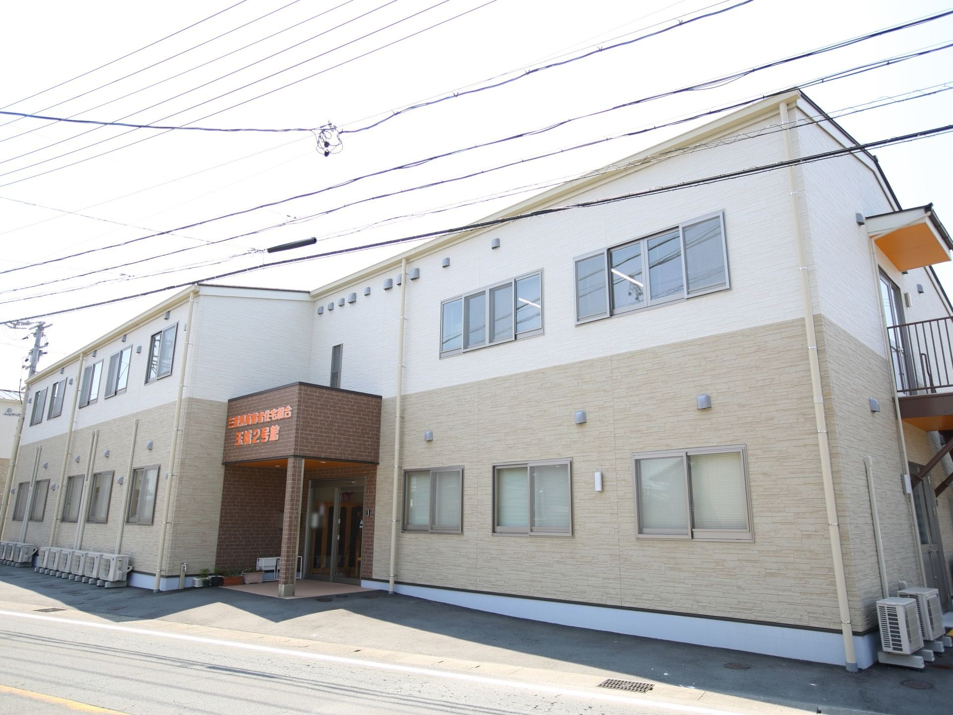 白とベージュのツートンカラーの外観である。２階建ての建物になっており、右手側には非常階段が設けられている。