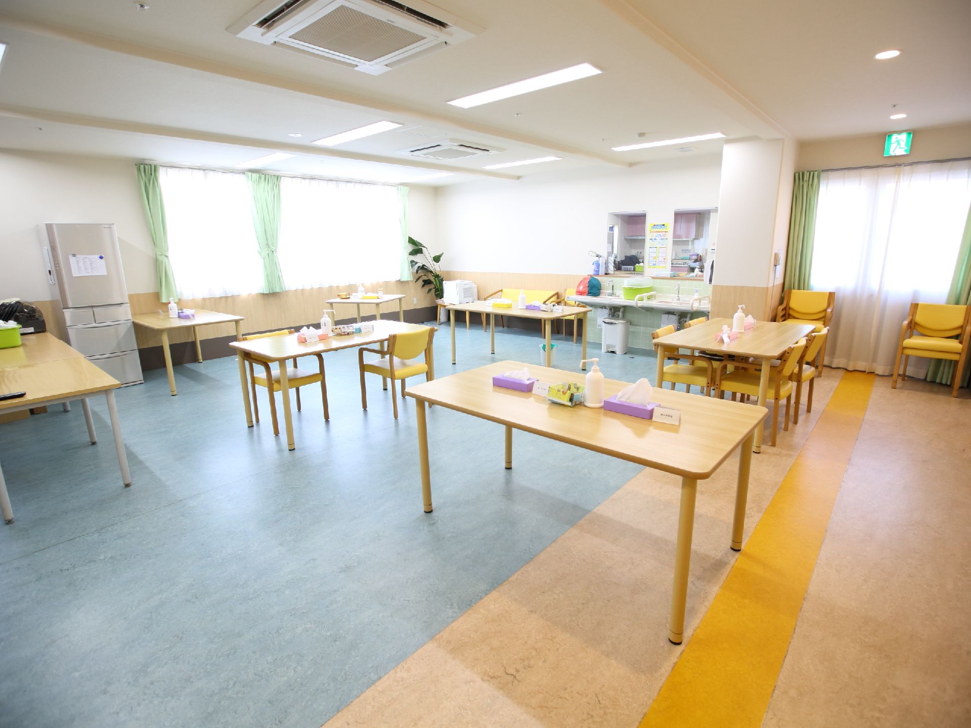 消毒用アルコールが置かれた複数のテーブルと、左手には冷蔵庫、奥には洗面台がある共有スペース