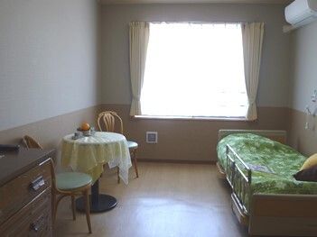 ベッドサイドには緊急通報装置があり、大きな窓にはカーテンが付いている居室には介護ベッドやテーブルセットがある。