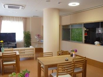 木製のダイニングテーブルが複数置かれ、日当たりのいい明るい食堂。花や観葉植物が飾られ、テレビが置かれている。