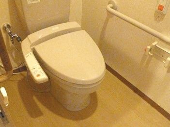 トイレはシンプルな設計で温水便座が付いている。壁には手すりやオレンジの呼び出しボタン、トイレットペーパーホルダーがある。