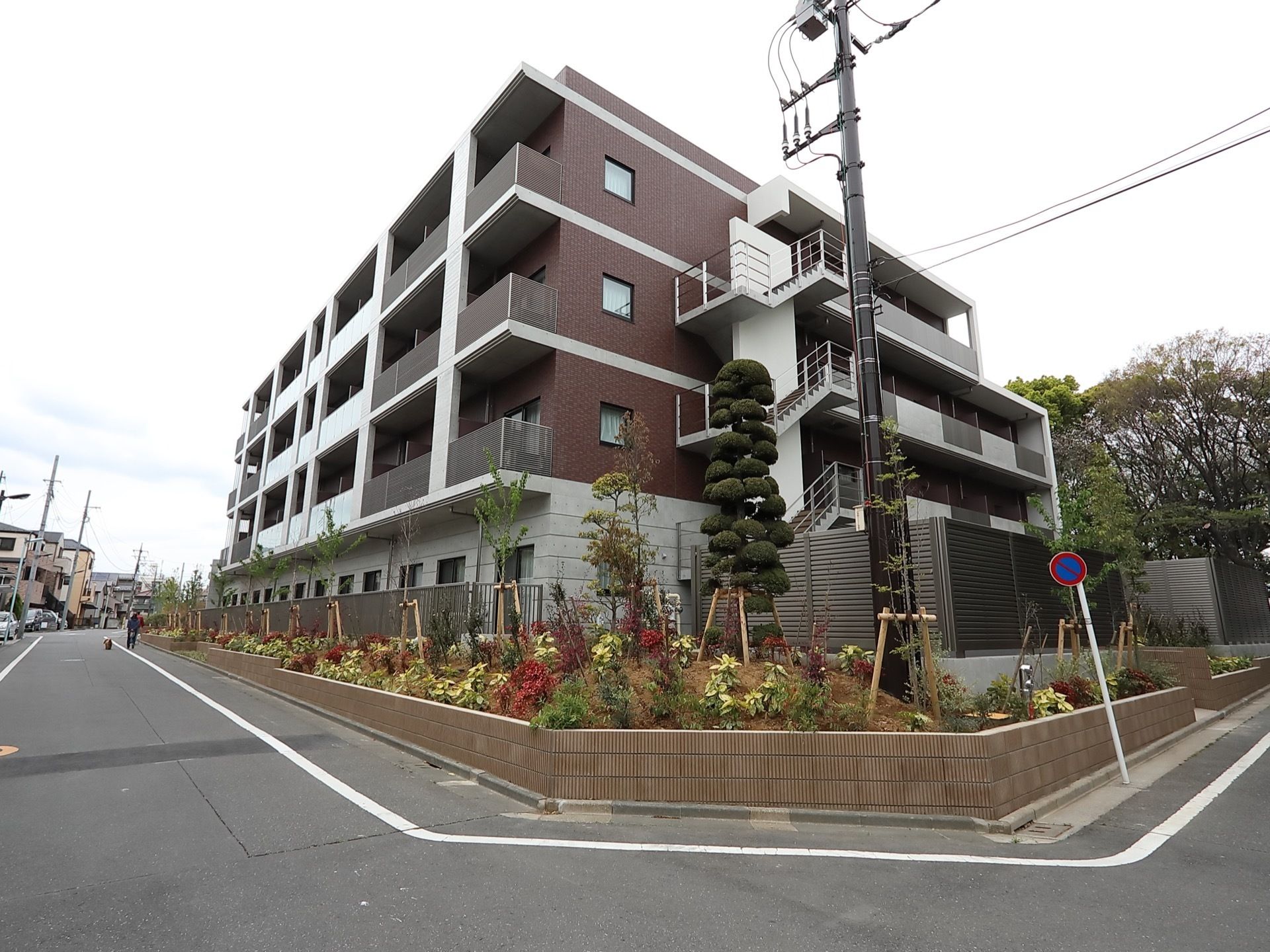 白と茶色の壁面の４階建ての大きな建物が角地になっている。周囲は草花が植えられている花壇があり、１階部分はスチールのフェンスで囲われている。