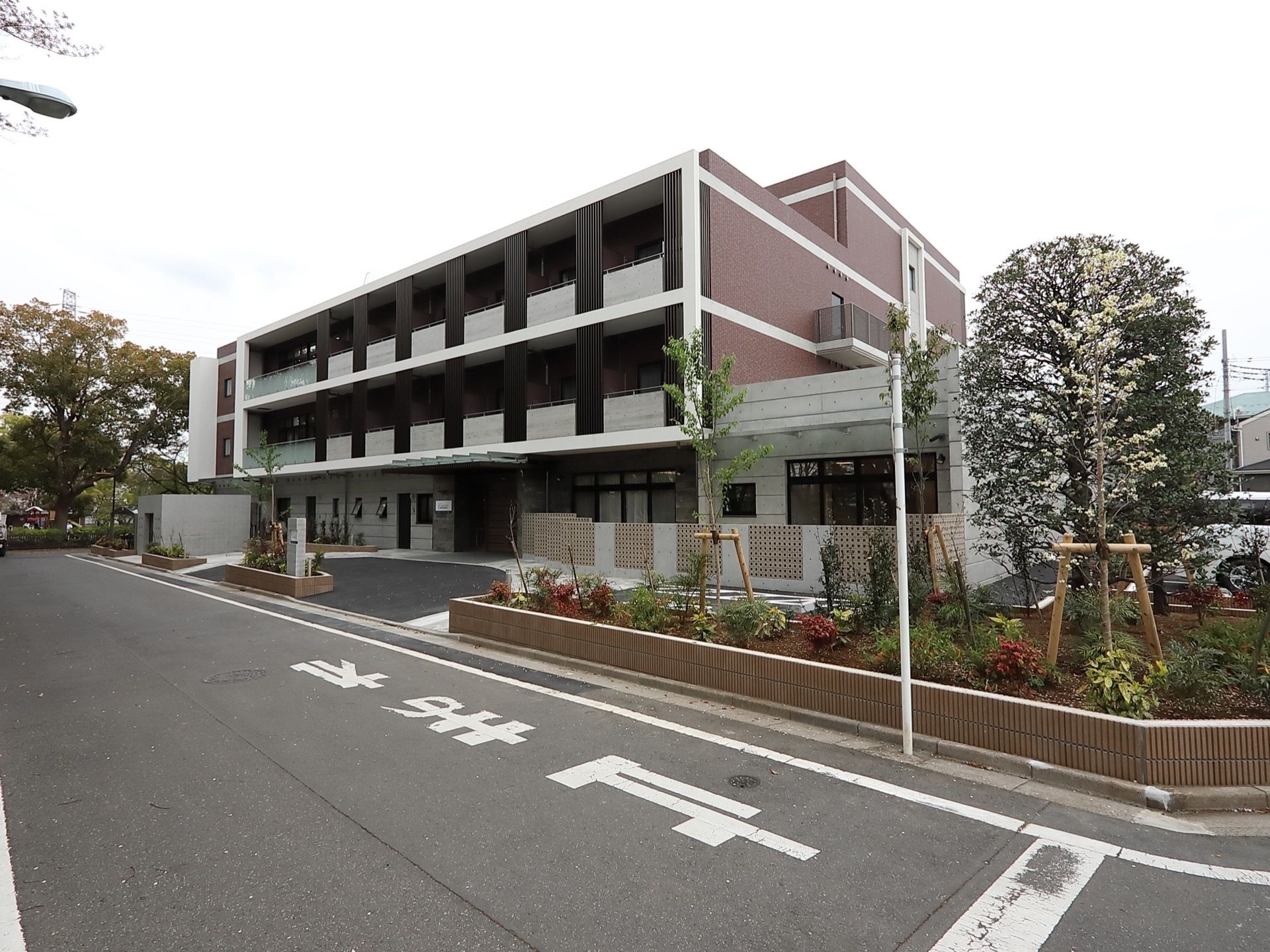 白と茶色の壁の四角い４階建ての建物がある。角地に建っており、周囲には大きな木や花壇があり、緑があふれている。