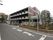 白と茶色の壁の四角い４階建ての建物がある。角地に建っており、周囲には大きな木や花壇があり、緑があふれている。
