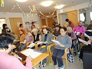 ホールに並べた椅子に座って歌詞カードを見ながら歌を歌う入居者の様子。