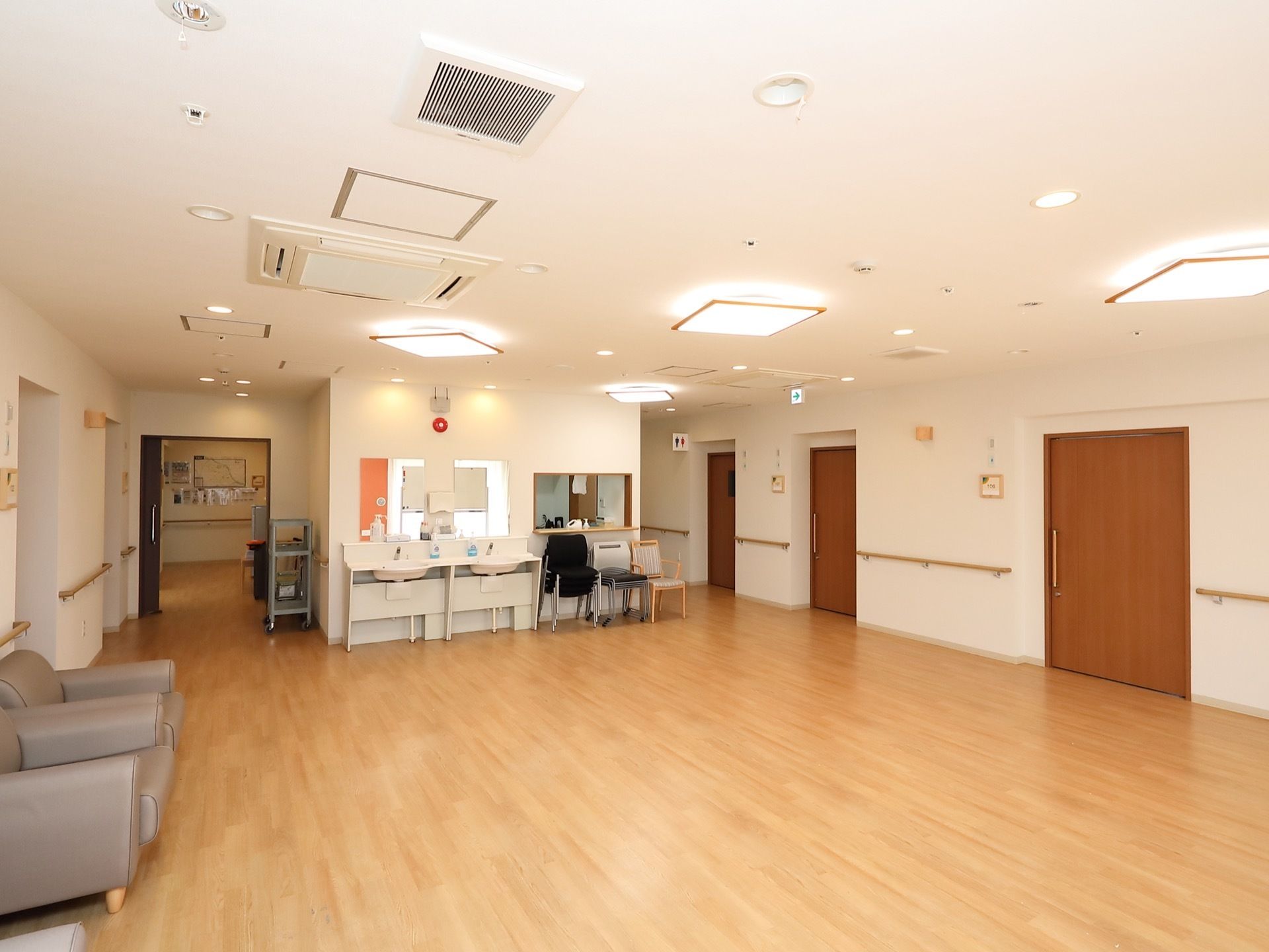 廊下から続く一角が広い空間になっており、奥の壁には洗面台が２か所にある。左手の壁際にグレーのソファが置かれている。