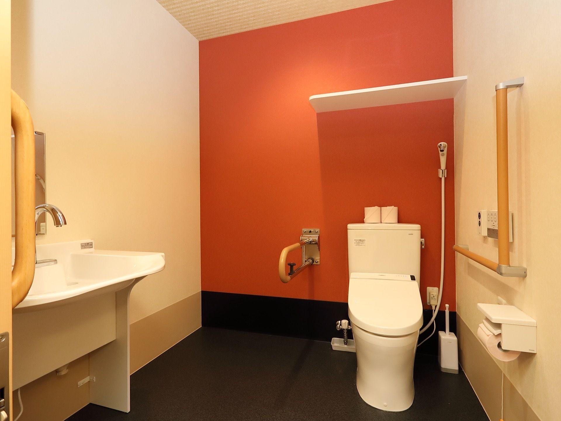 トイレの壁の一面は赤色になっている。便器洗浄用のシャワーが設置されている。自動水栓の洗面台が入口近くにある。