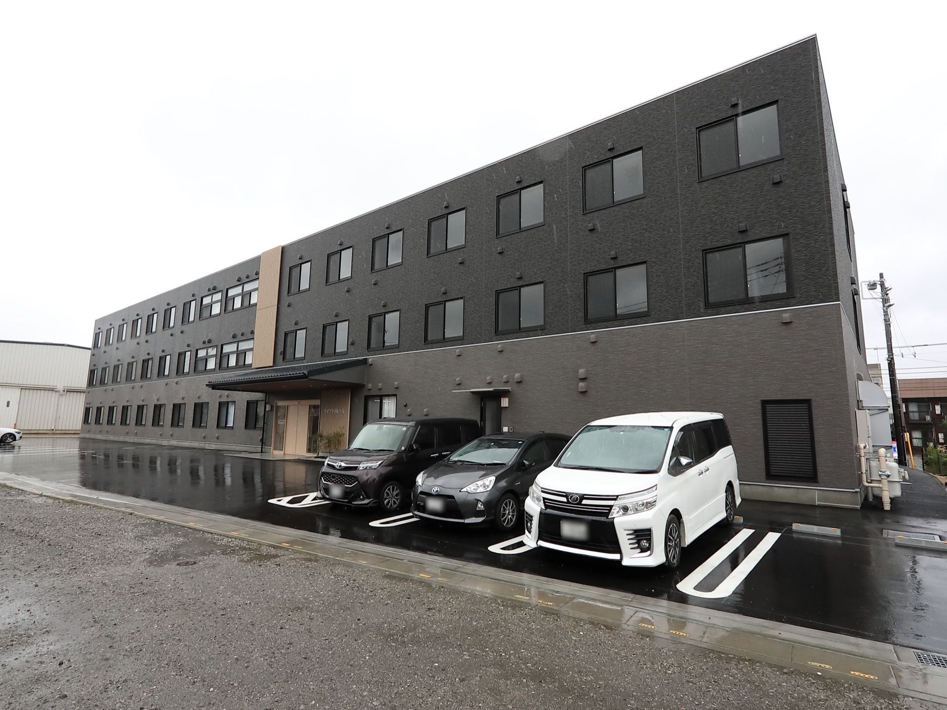 ダークブラウンでまとめられた落ち着いた雰囲気の3階建ての建物。駐車スペースもある。
