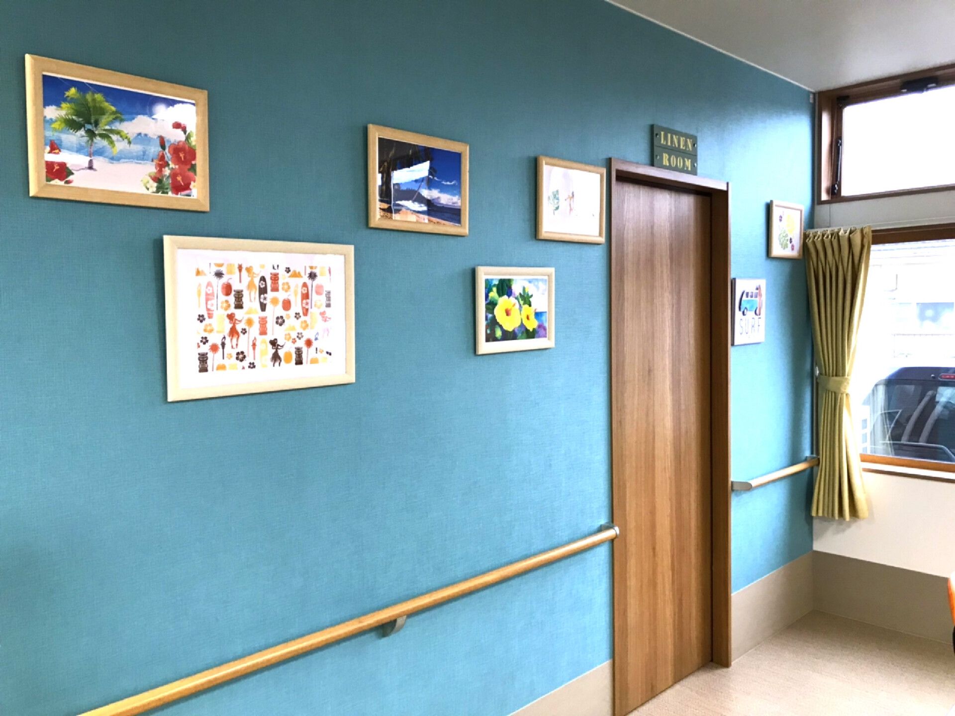 ハイビスカスの花やビーチの絵画が飾られた廊下の壁には木製の手すりも付いている。落ち着いたブルー系の壁紙は海のような色合い。