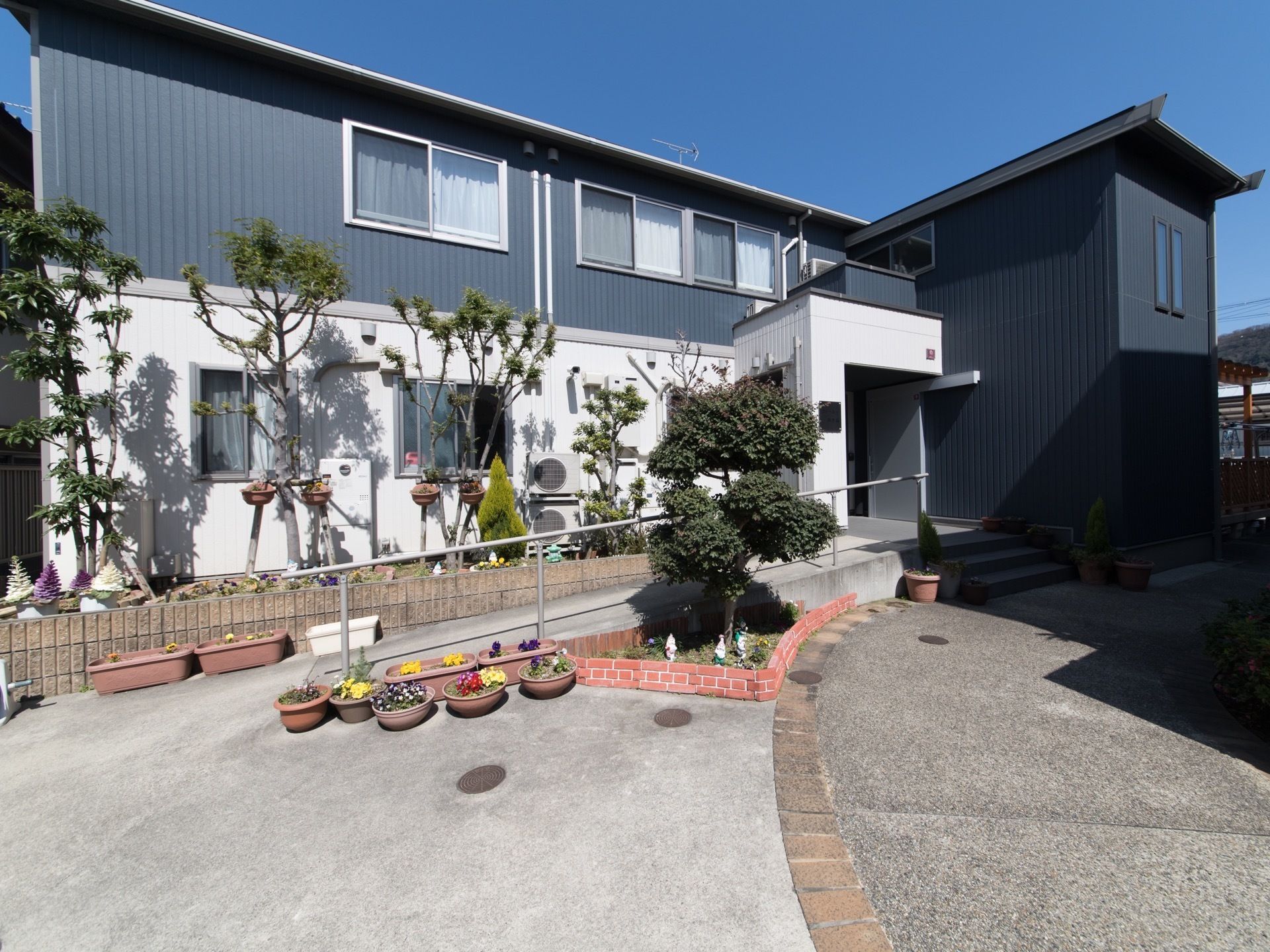 地上２階建て。玄関の前には階段と手すり付きのスロープが設置されている。色とりどりの花や木が植えられている。