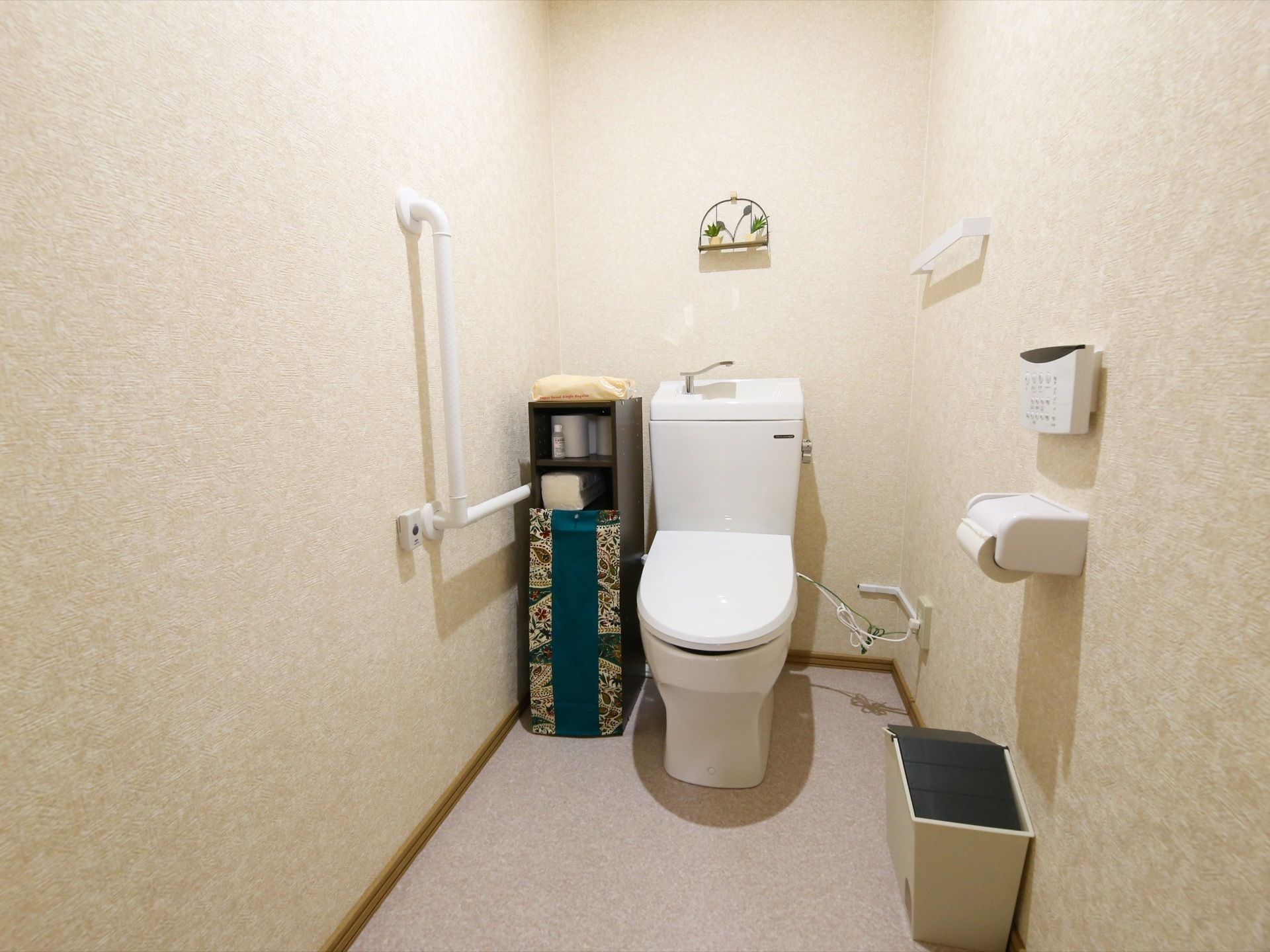 壁にL字型手すりが設置され、温水洗浄便座器が完備されたトイレである。手すりと便座器の間に収納ボックスが置かれている。