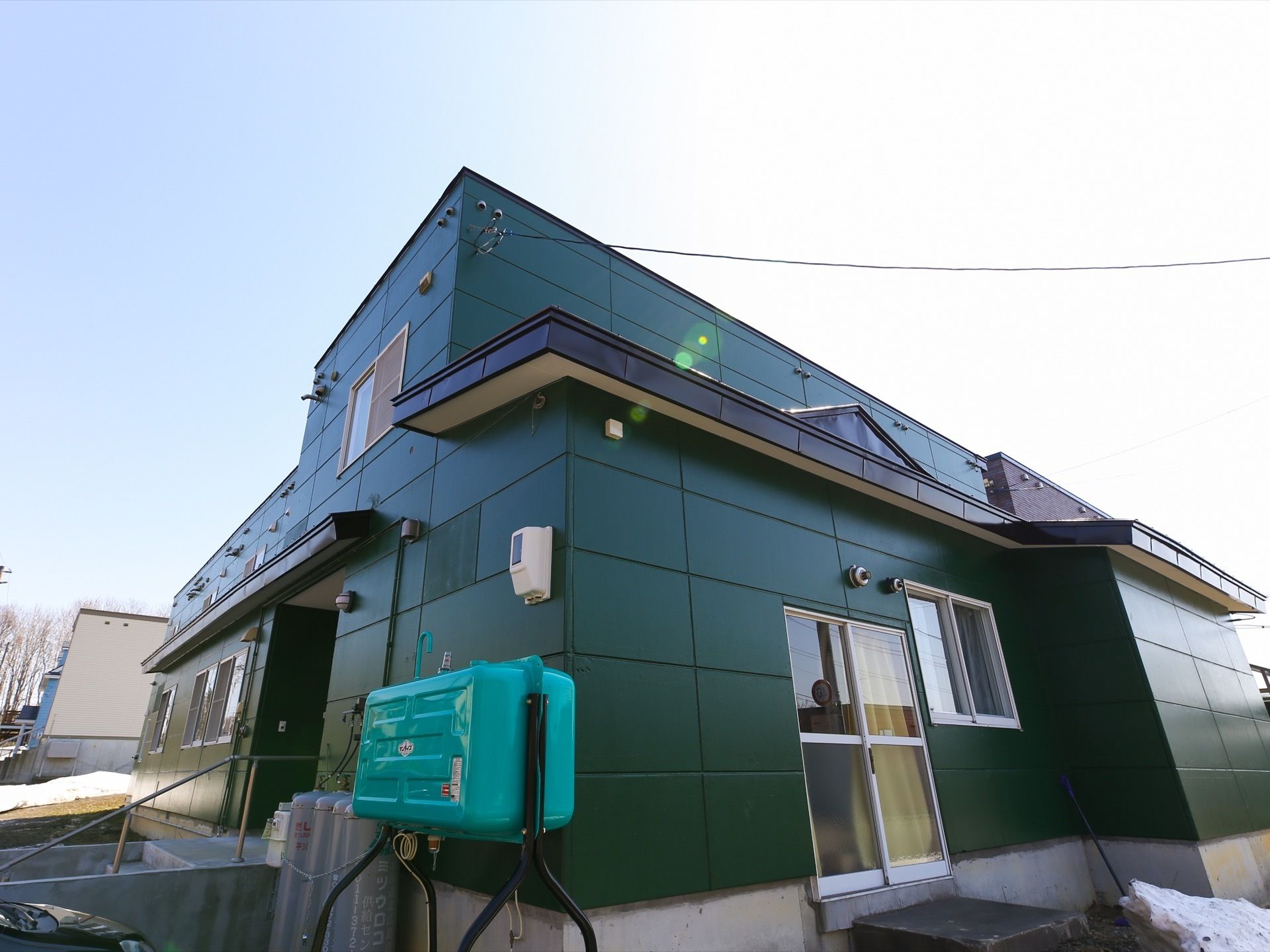 降り積もった雪が残る晴れた日の２階建て施設の外観である。濃いグリーンの外壁で、プロパンガスが外壁に沿って並べられている。