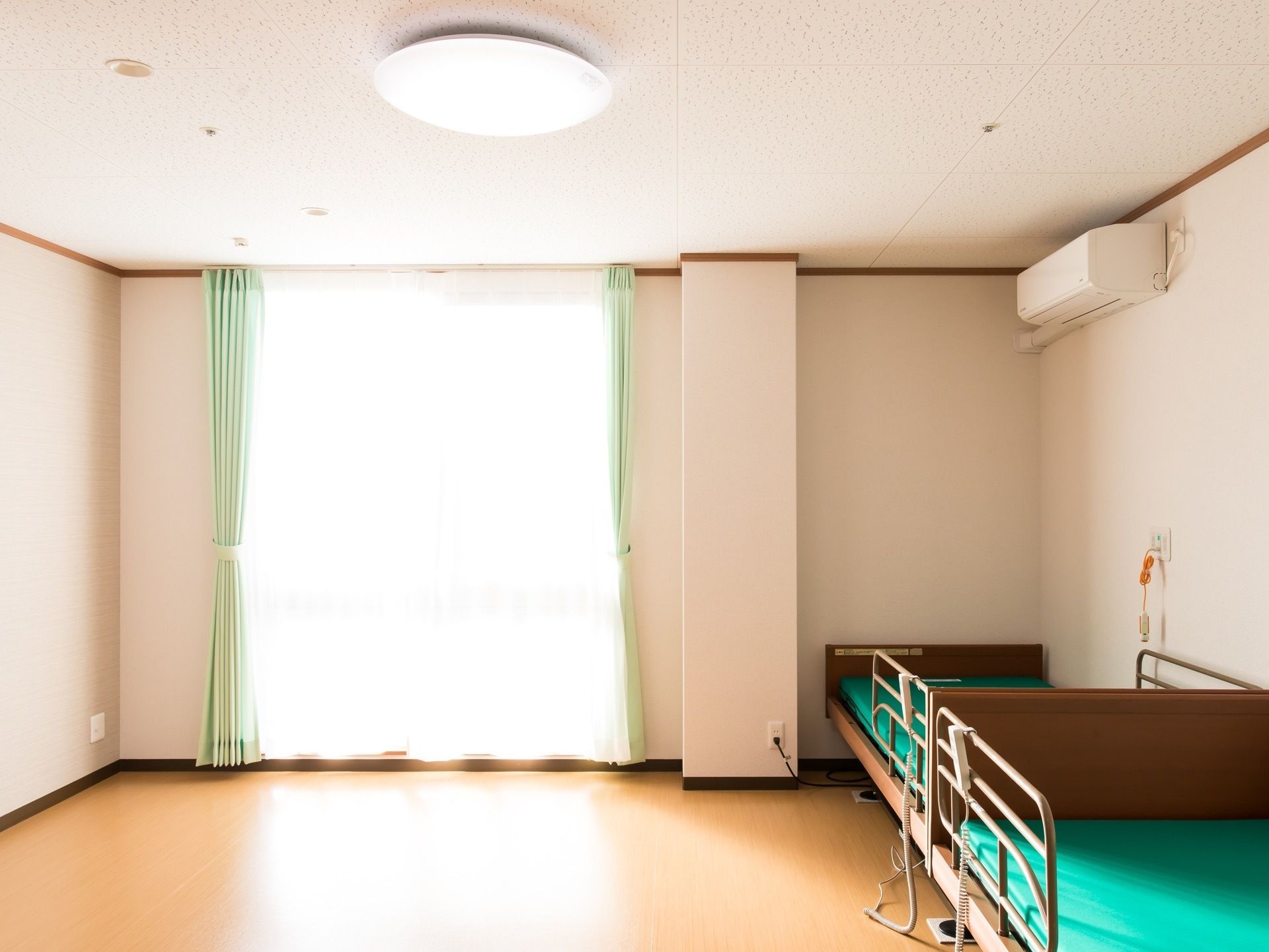 居室の壁際に、転落防止柵がついた介護ベッドが２台並べて置かれている。その上に白色のエアコンを設置。