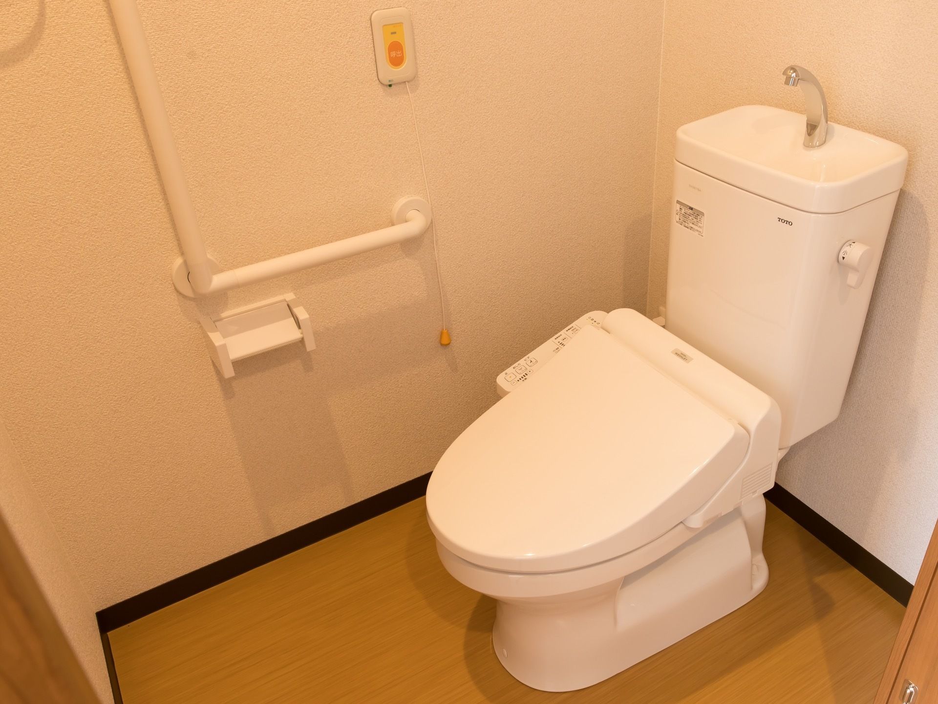 引き戸を開けた所にトイレがある。便座横の壁に、オレンジ色の呼び出しボタンがあり、ひもがぶら下がっている。