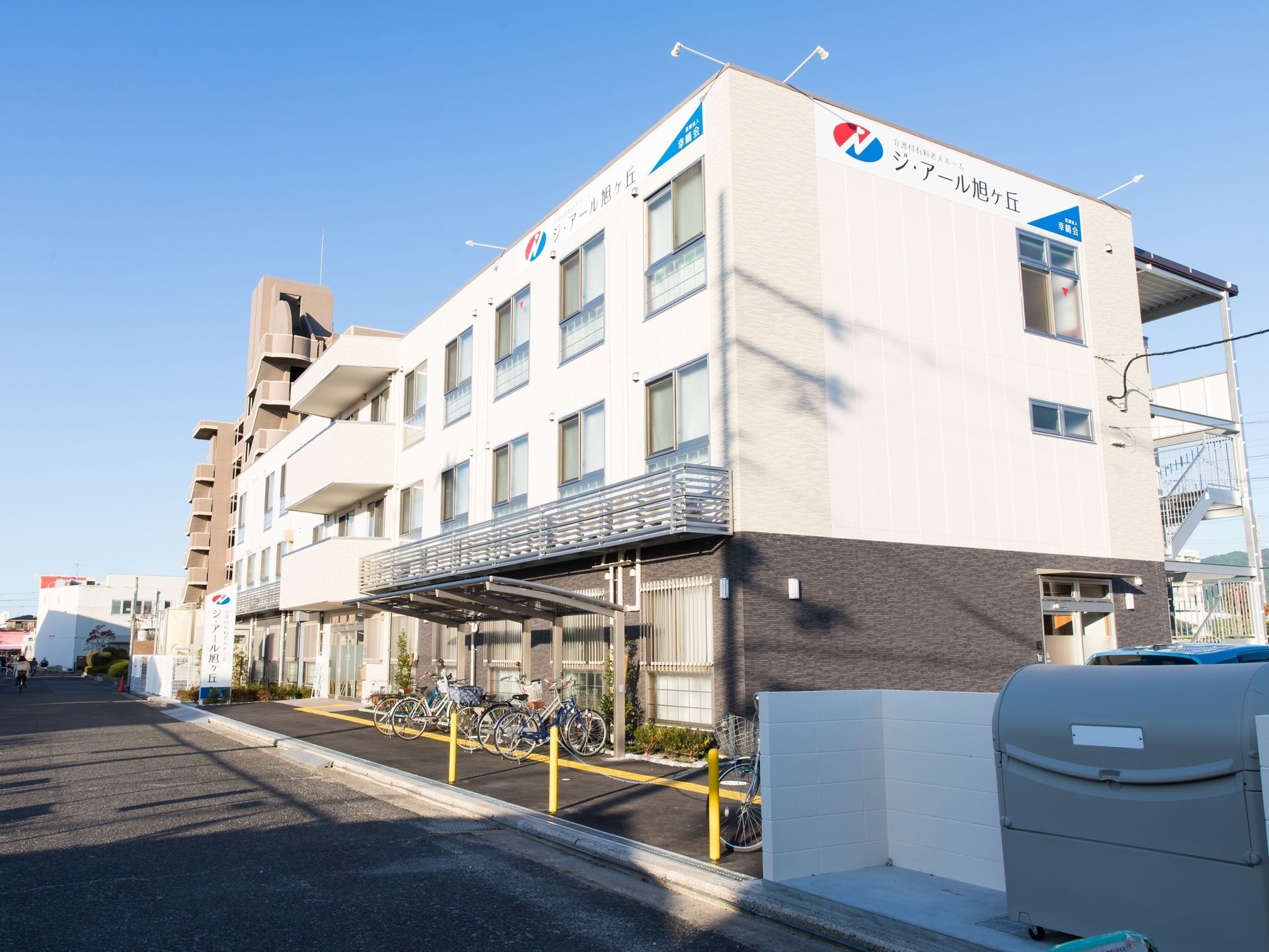 道路に面して建っている３階建ての施設である。玄関横にカーポートが設置され、その下に多くの自転車が停まっている。