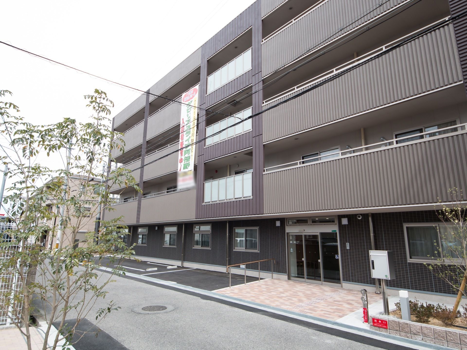 シックな色合いに施された4階建ての建物である。入り口付近には駐車スペースが設けられている。外壁には施設名が記された広告が設置されている。