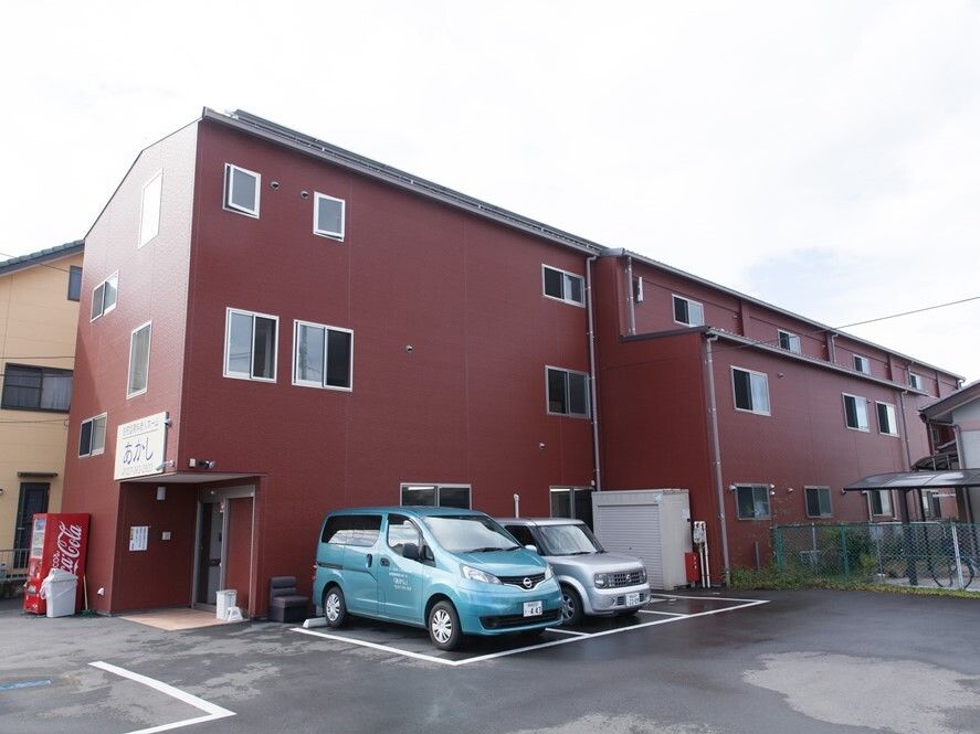 ブラウン系の3階建ての建物の玄関の上にはホーム名が書かれた看板があり、広い駐車場も付いている。