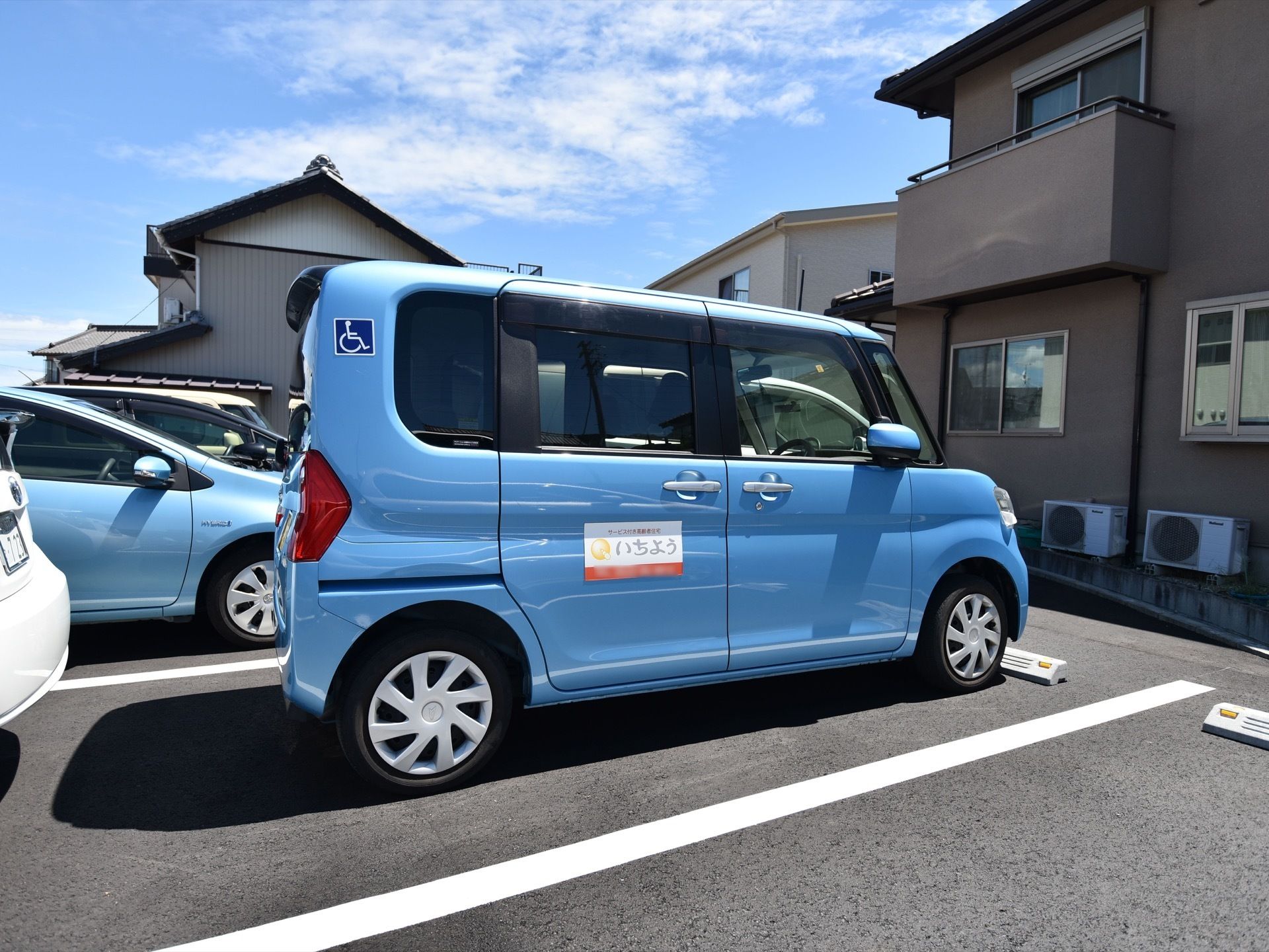 エントランスの近くに駐車スペースが確保され、施設の送迎車が停められている。小回りの利く小ぶりの車体に施設名の入ったステッカーが貼られている。