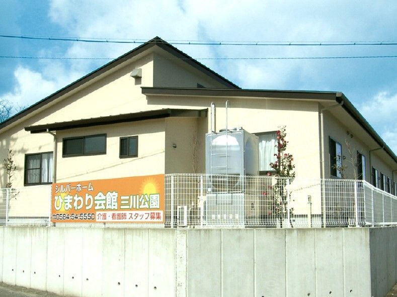 平屋建ての建物はベージュの外壁で建物の周りには植物も植えられ、看板が付いたフェンスも建てられている。