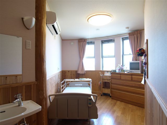 介護用ベッドとたんす、テレビがある施設内の居室。手前に洗面台がついている写真