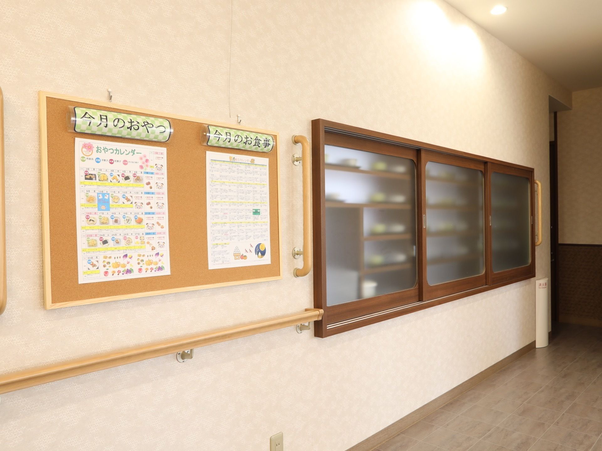 今月のお食事とおやつのメニューが食堂に掲示板に貼り出されている。隣にはできあがった食事がトレーに置かれている。