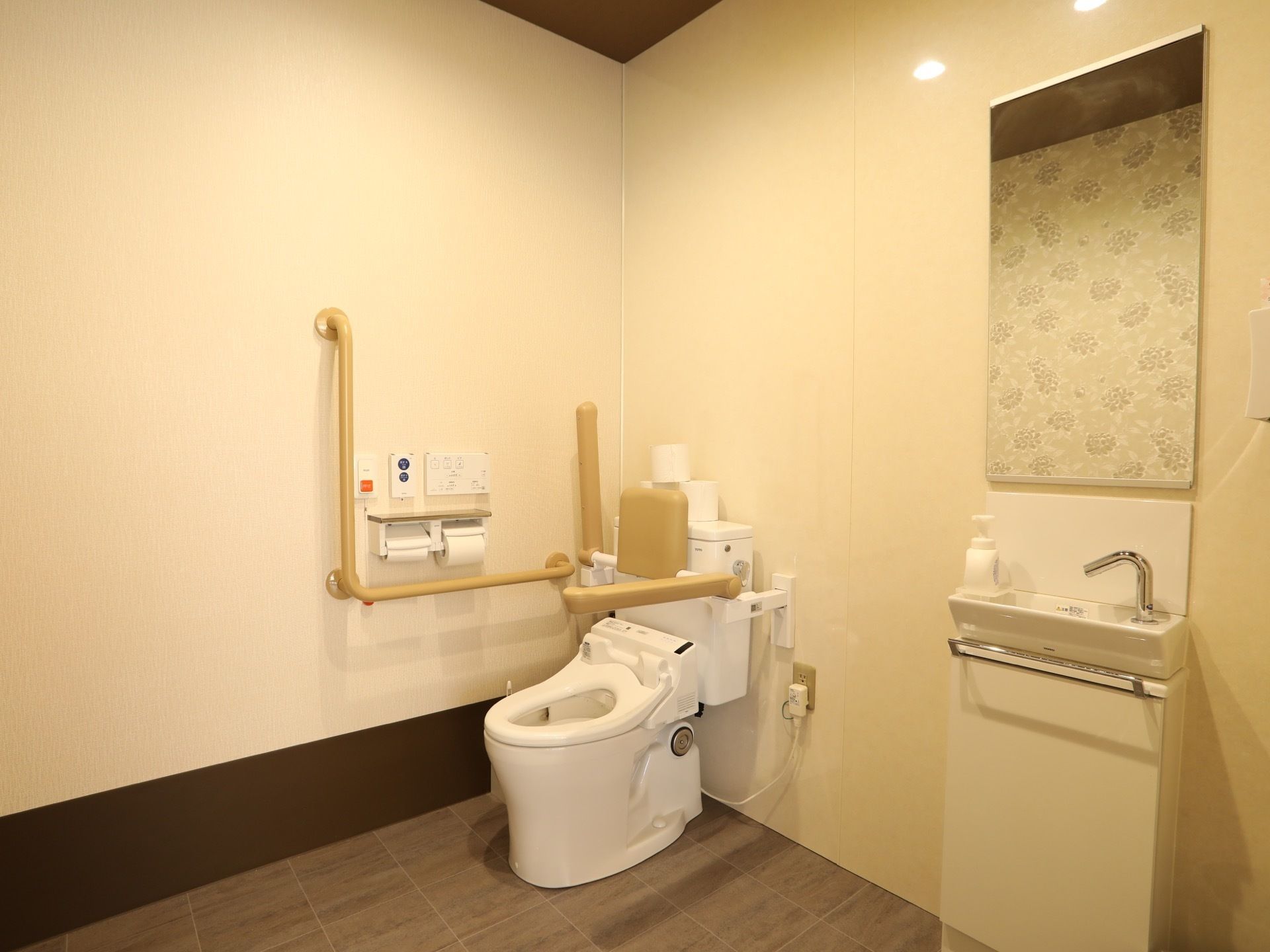 広めの空間には、トイレのほかにミラーのついた手洗器が置かれている。ベージュ色の壁で、一部に使用されている花柄の壁紙が印象的である。