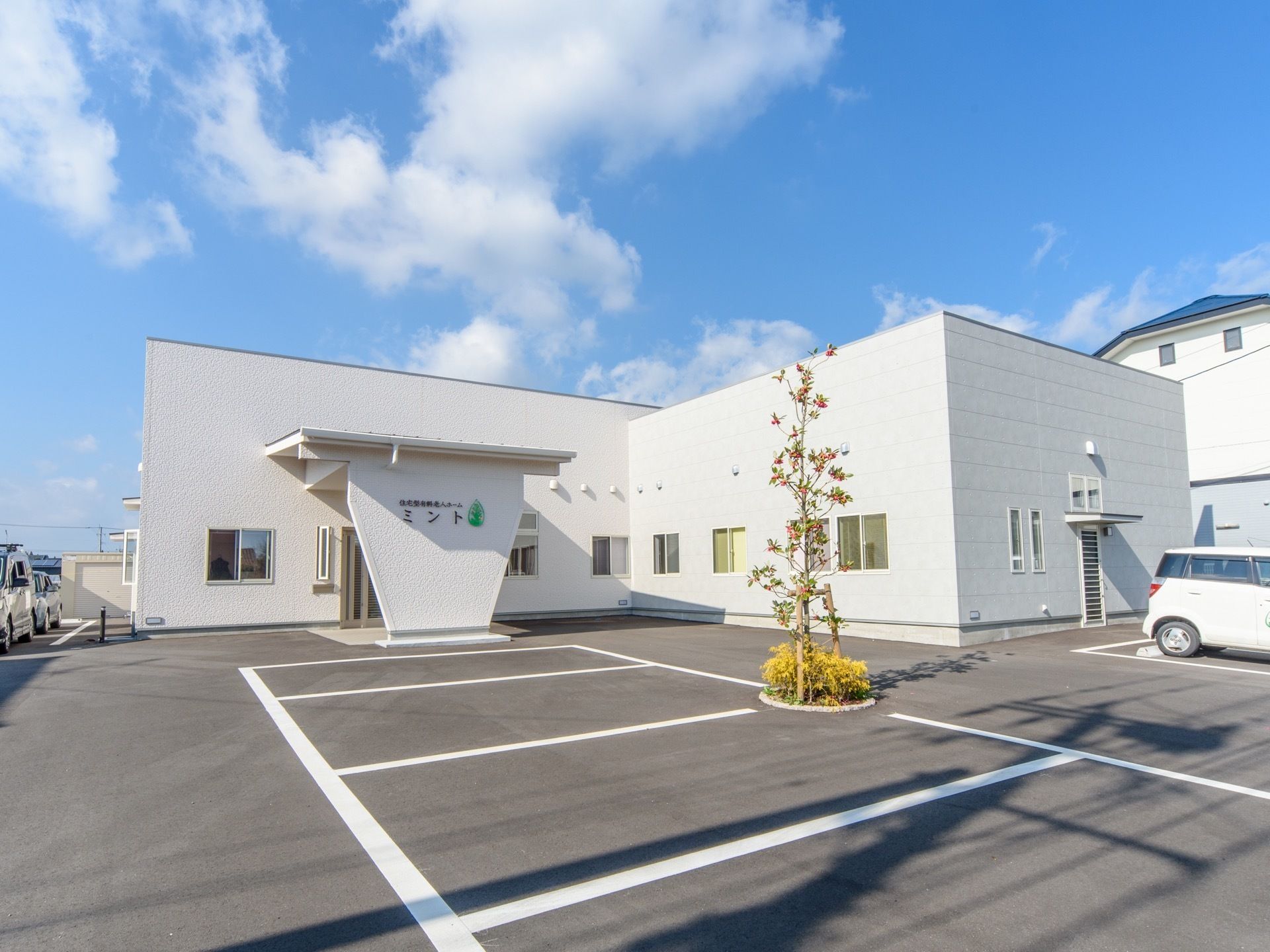中央奥に白い施設が建っている。建物の前は駐車スペースが広がっており、樹木が１本植えられている。左奥に車が１台停まっている。