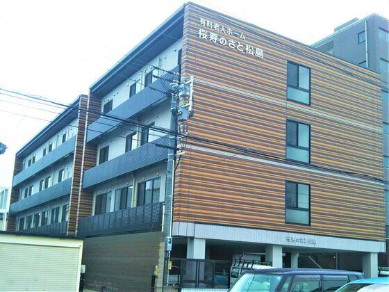 ブラウン系の４階建ての建物。周辺にも背の高い建物があり、駐車場も完備されている。