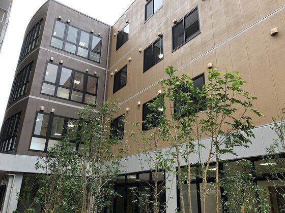 ４階建ての建物はブラウン系でまとめられ、明るい雰囲気。建物の周辺には木がたくさん植えられている。