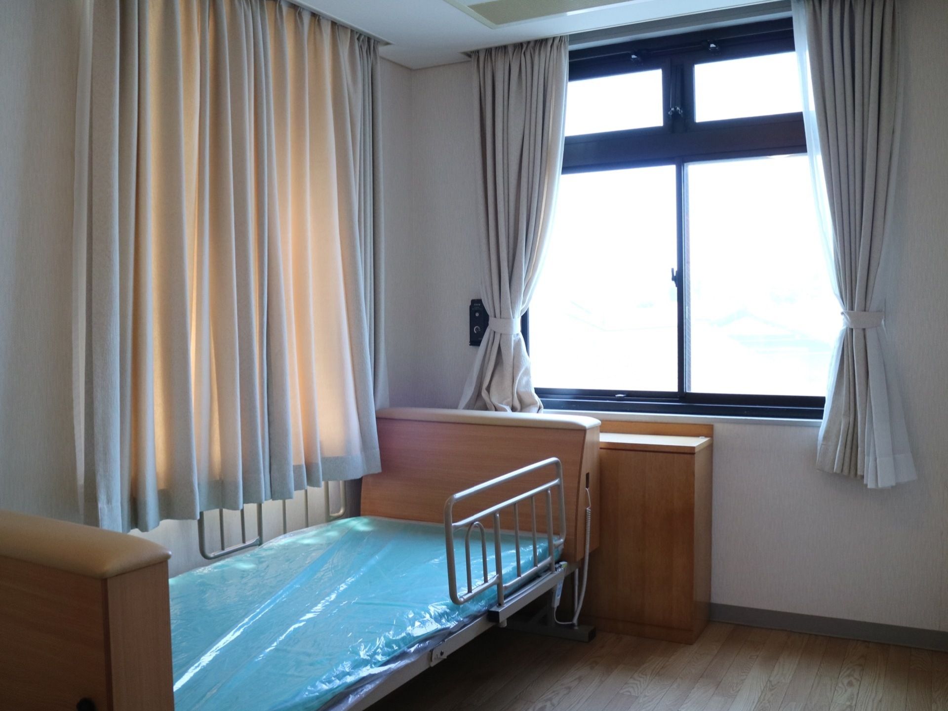 居室は個室になっている。２箇所に設けられた窓にはカーテンが掛かっており、介護ベッドやナースコールが完備されている。