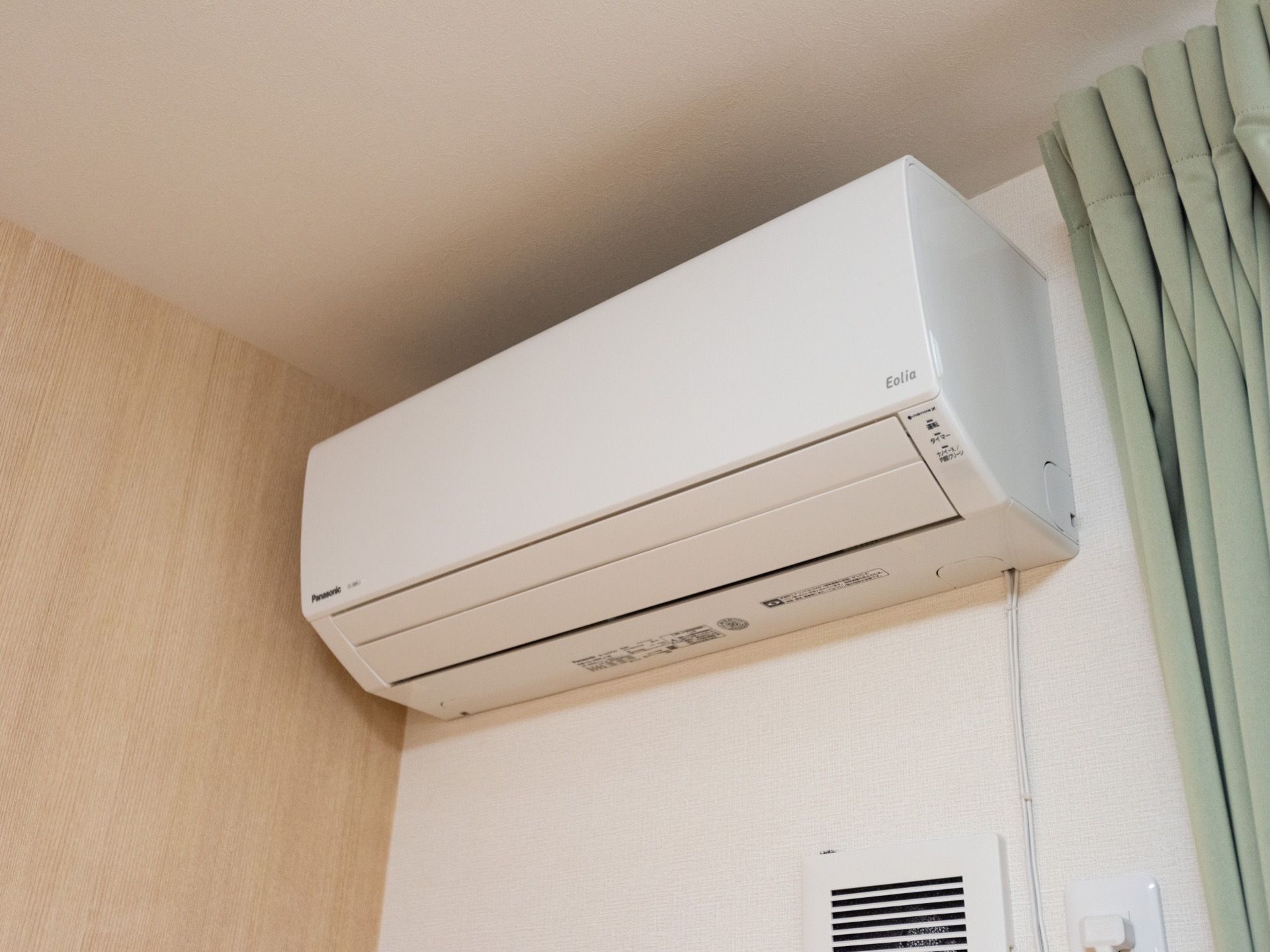白い壁と木目調の壁があり、白い壁の方に大きなエアコンが取り付けられている。右側に緑のカーテンが掛けられている。