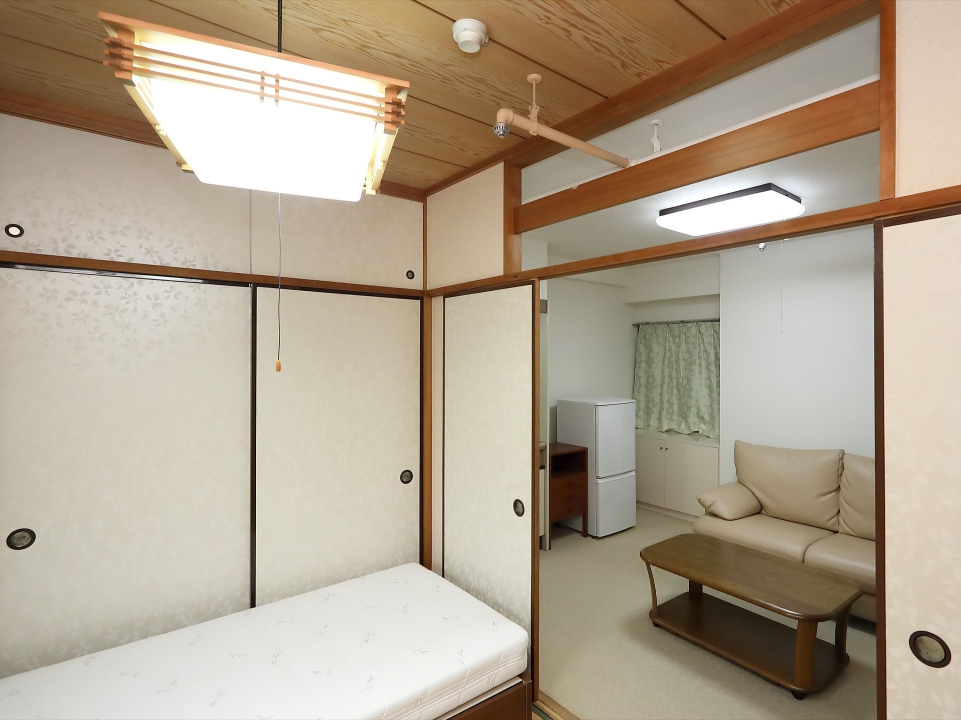 和室の一角に寝具が据えられている。和室から出たところに、ソファとテーブルが置かれており、奧には冷蔵庫が見える。