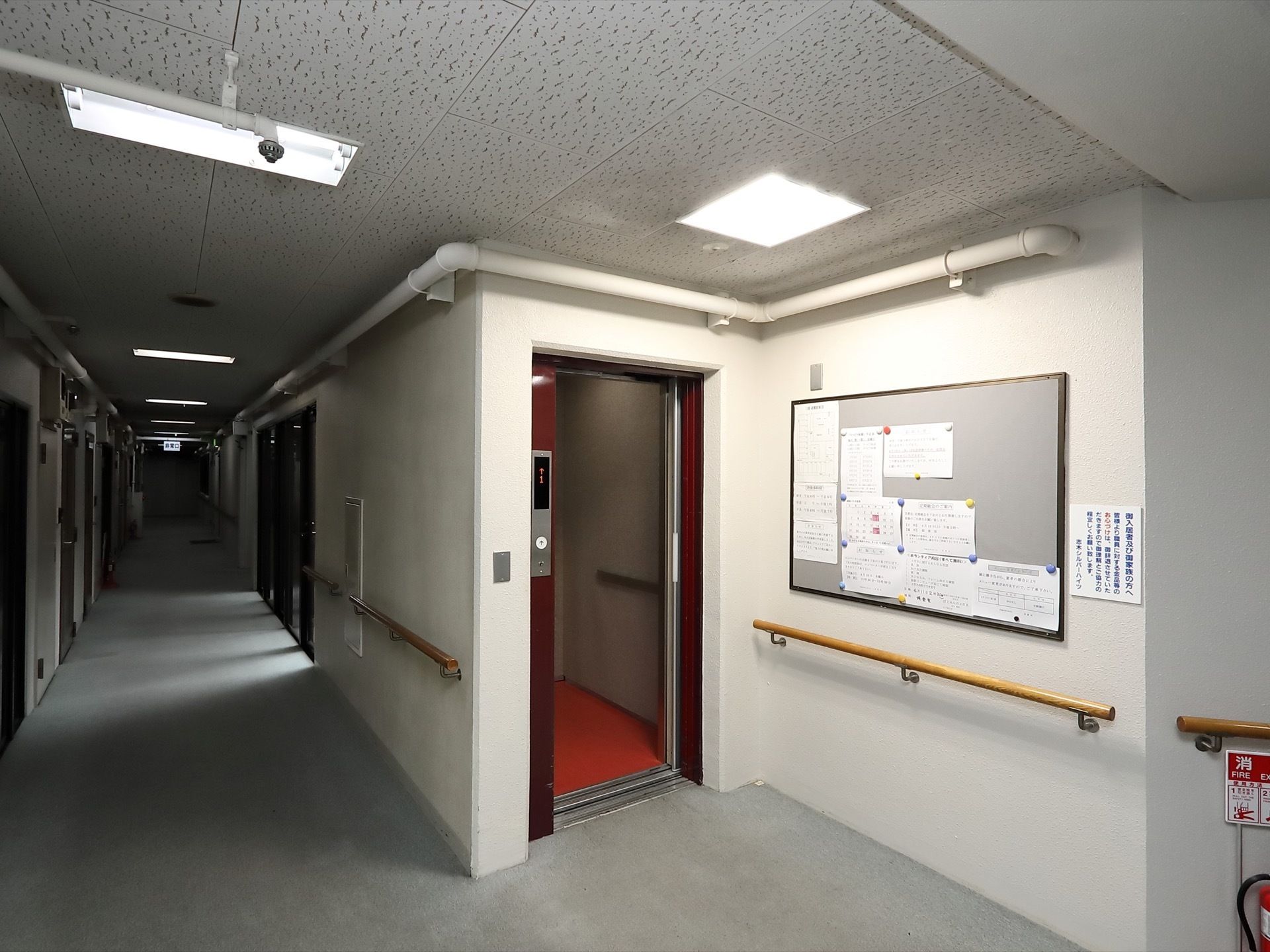 エレベーター前の天井には大きな照明があり、左側の通路よりも明るい。右側の壁面には掲示板や手すりが設置されている。