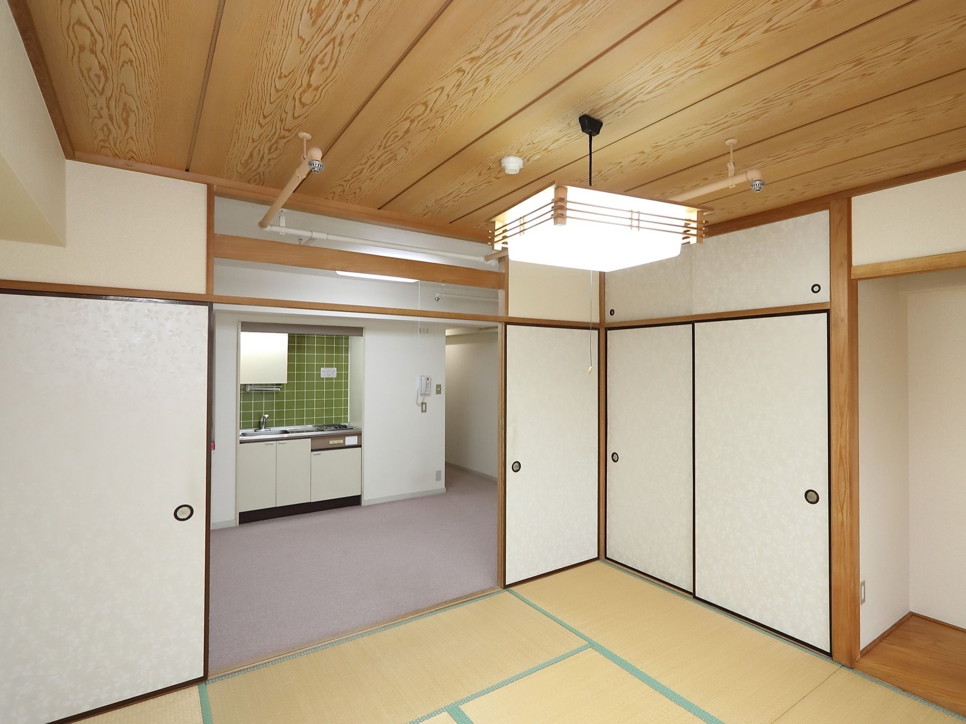 和室の中央に、大きな照明が提げられている。和室の向こう側には、壁面にタイルが貼られたキッチンスペースが見える。