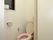 壁の高い位置に窓が設置されており、予備のトイレットペーパーが２つ置いてある。ピンク色の便座カバーとマットが取りつけられている。