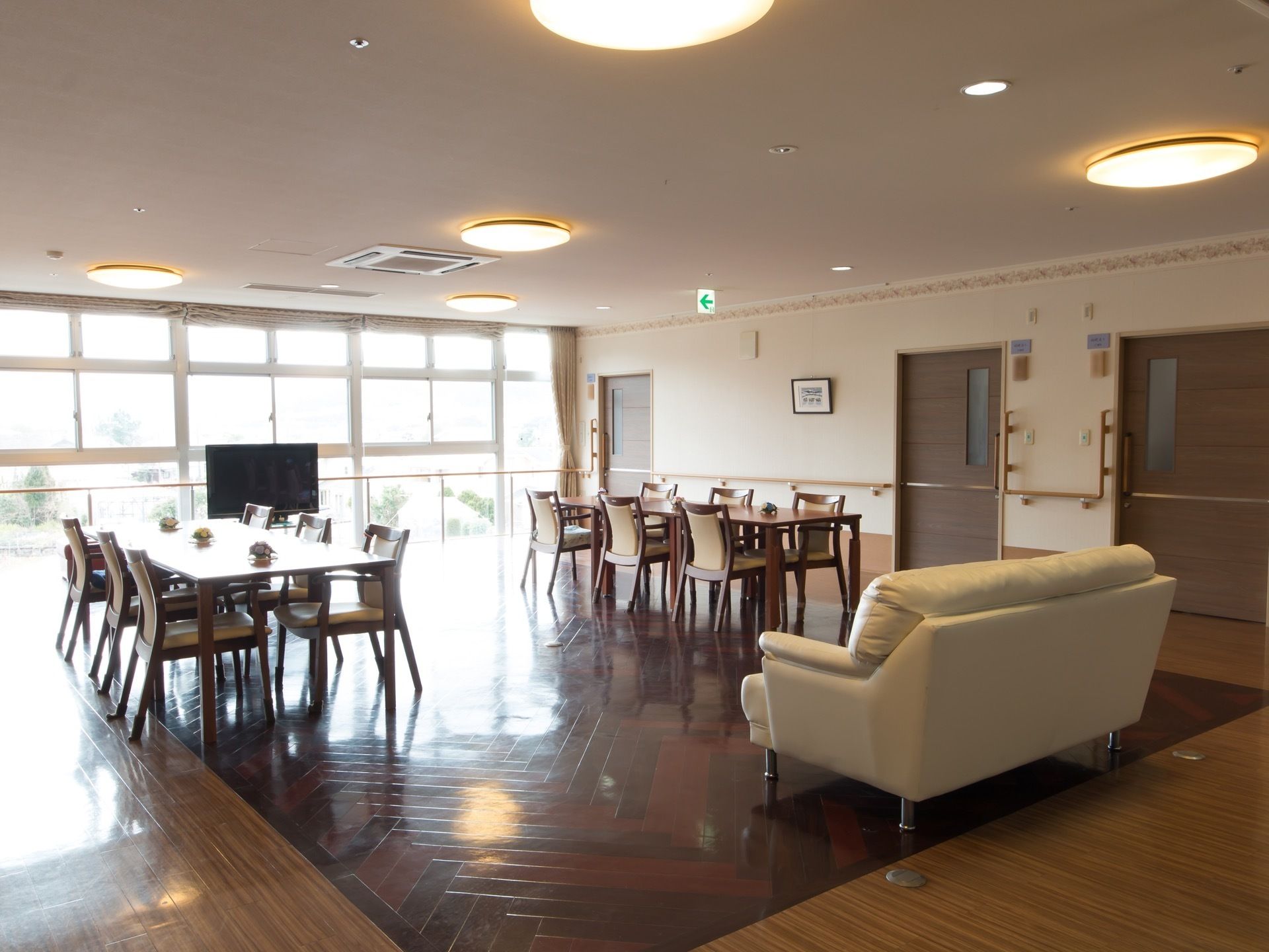 窓の広い部屋である。手前にソファーが置かれている。ダイニングテーブルと椅子が、縦に２列並んでいる。床の木目がきれいである。