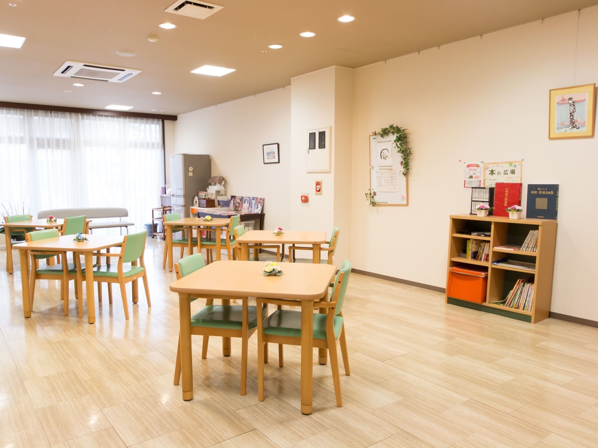 窓からの陽光で明るい部屋である。木製のテーブルと椅子がセットされ、数組置かれている。部屋の中央に本棚があり、数冊の本が収納されている。
