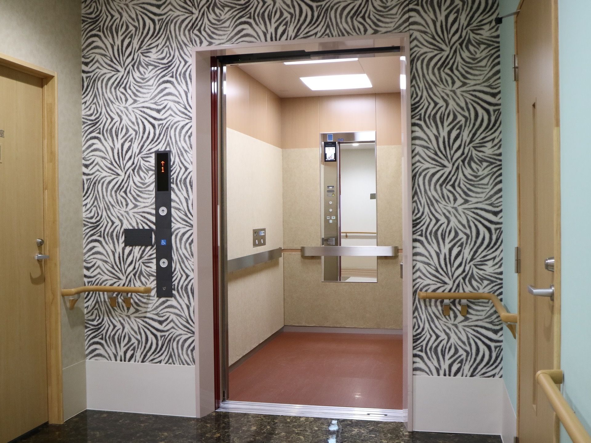 エレベーターの扉は開いており、中の様子が見える。床は赤色で背面には縦長の鏡が設置されている。手すりもある。