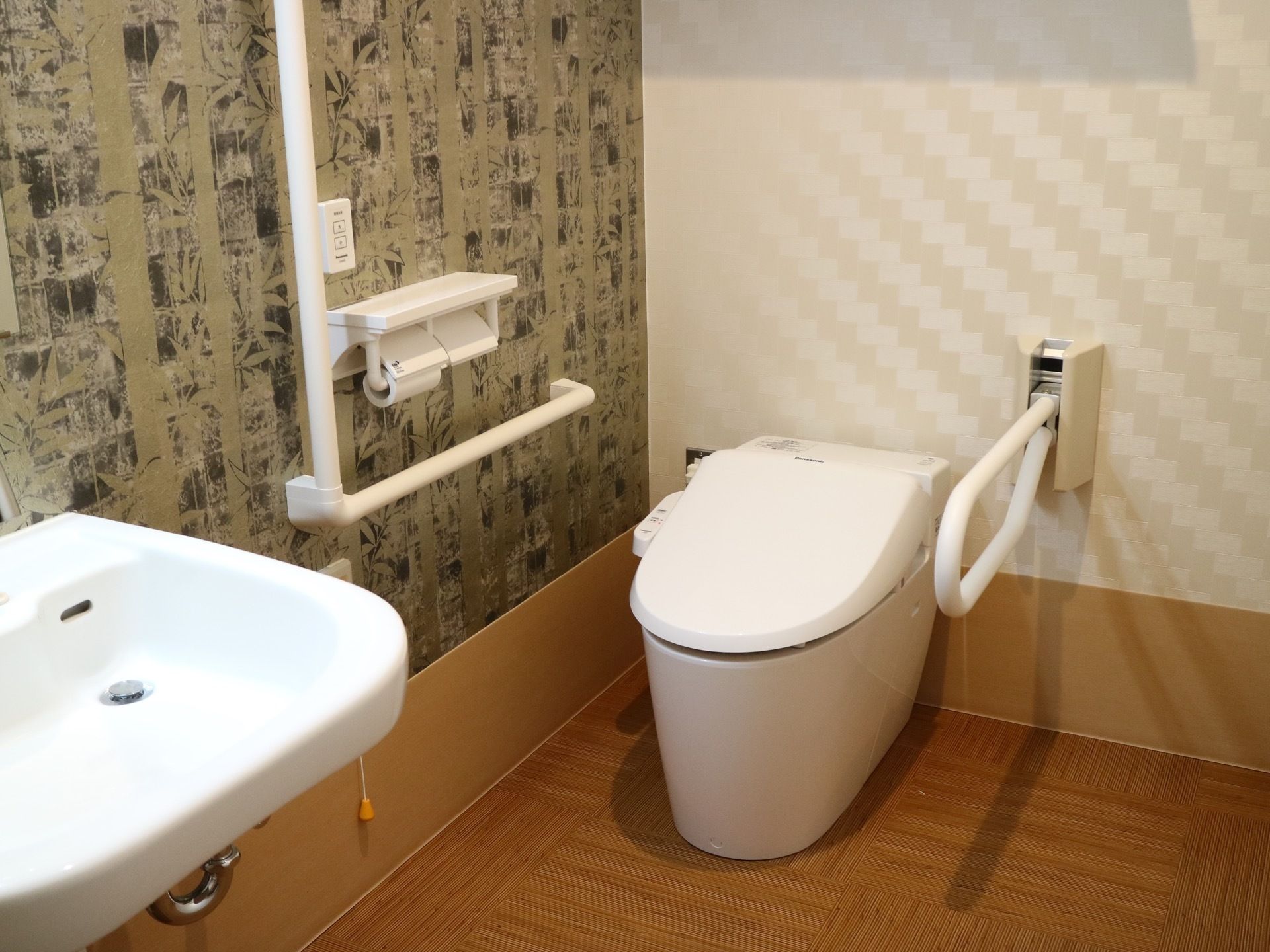 壁の一部が柄の付いたオリーブグリーンの壁紙のトイレがある。壁には手すりと可動式のひじ掛けが付いており、側には洗面台がある。