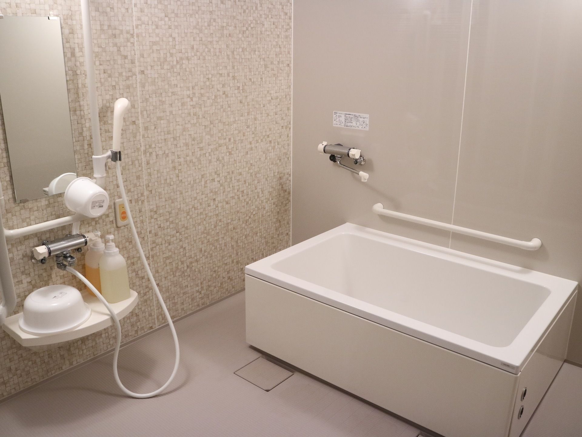 グレーとモザイク調の壁紙の浴室がある。浴槽の側の壁には手すりが設置されている。洗い場には洗面道具が置かれている。
