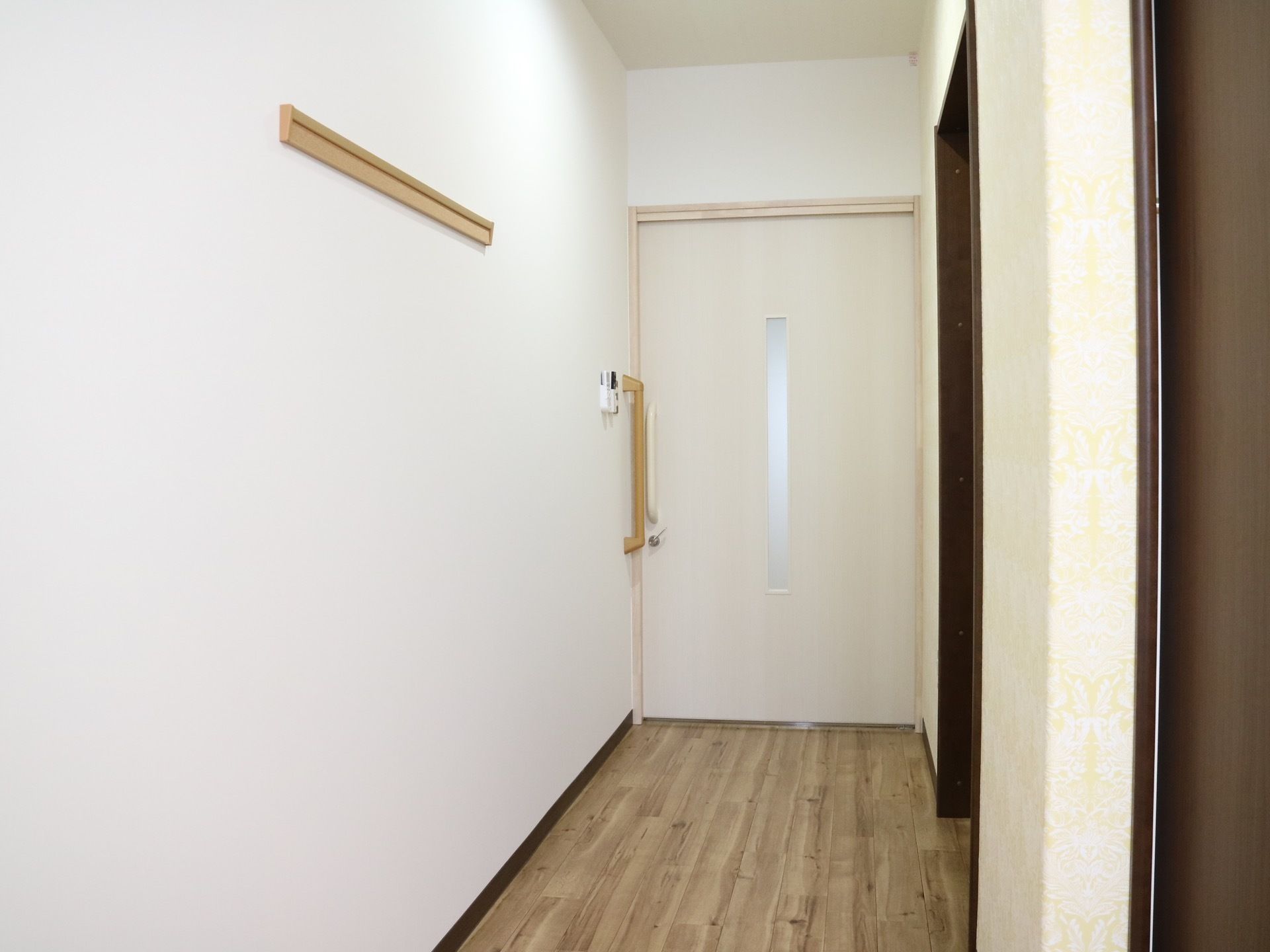 木目調の床の居室がある。壁と引き戸のドアは白く、ドアには小窓が付いている。入口付近には手すりが設置されている。