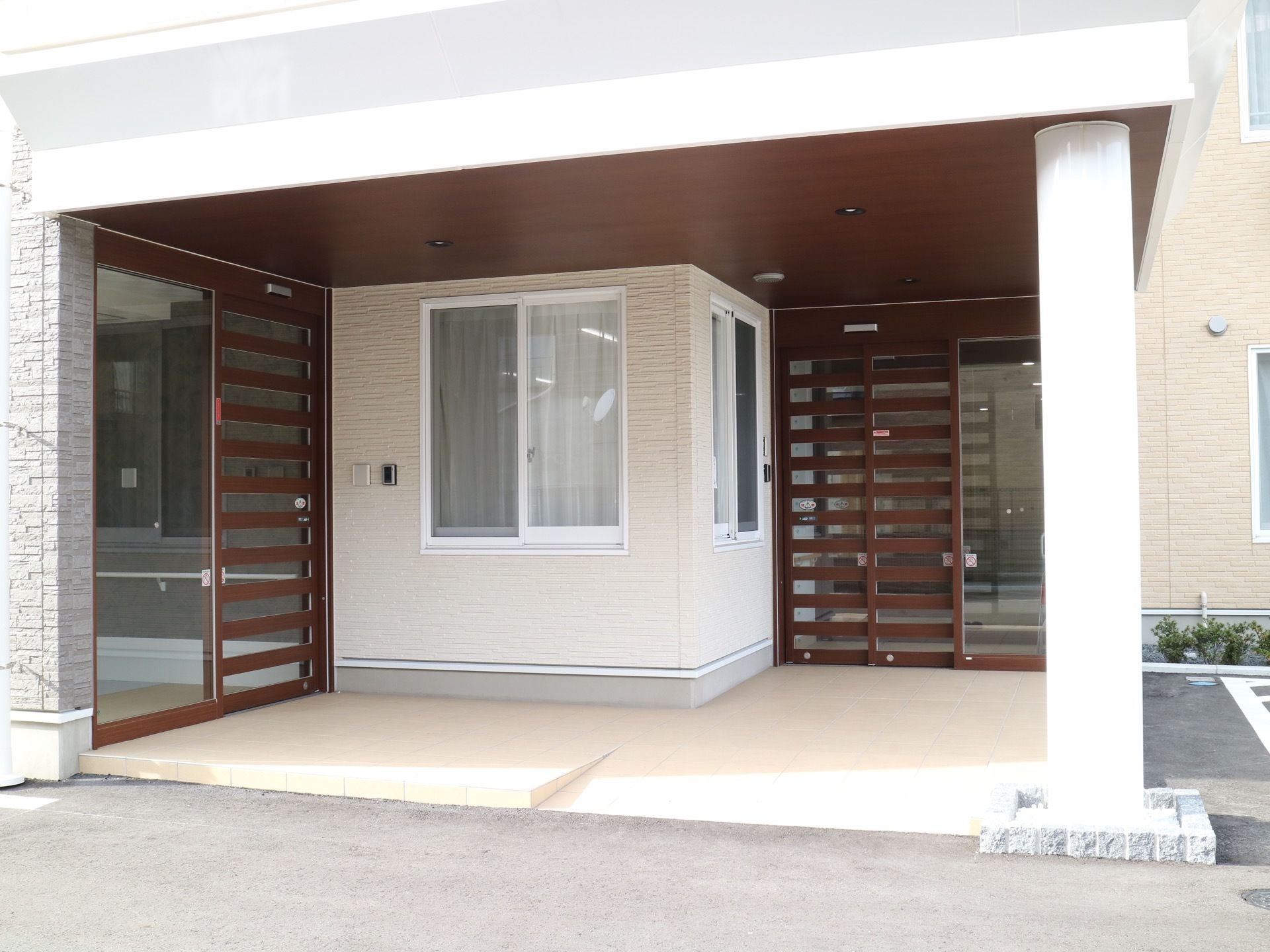 玄関の上に大きなルーフが設置されている。ガラス扉とダークブラウンの格子がはまった玄関ドアがある。足元には段差がない。