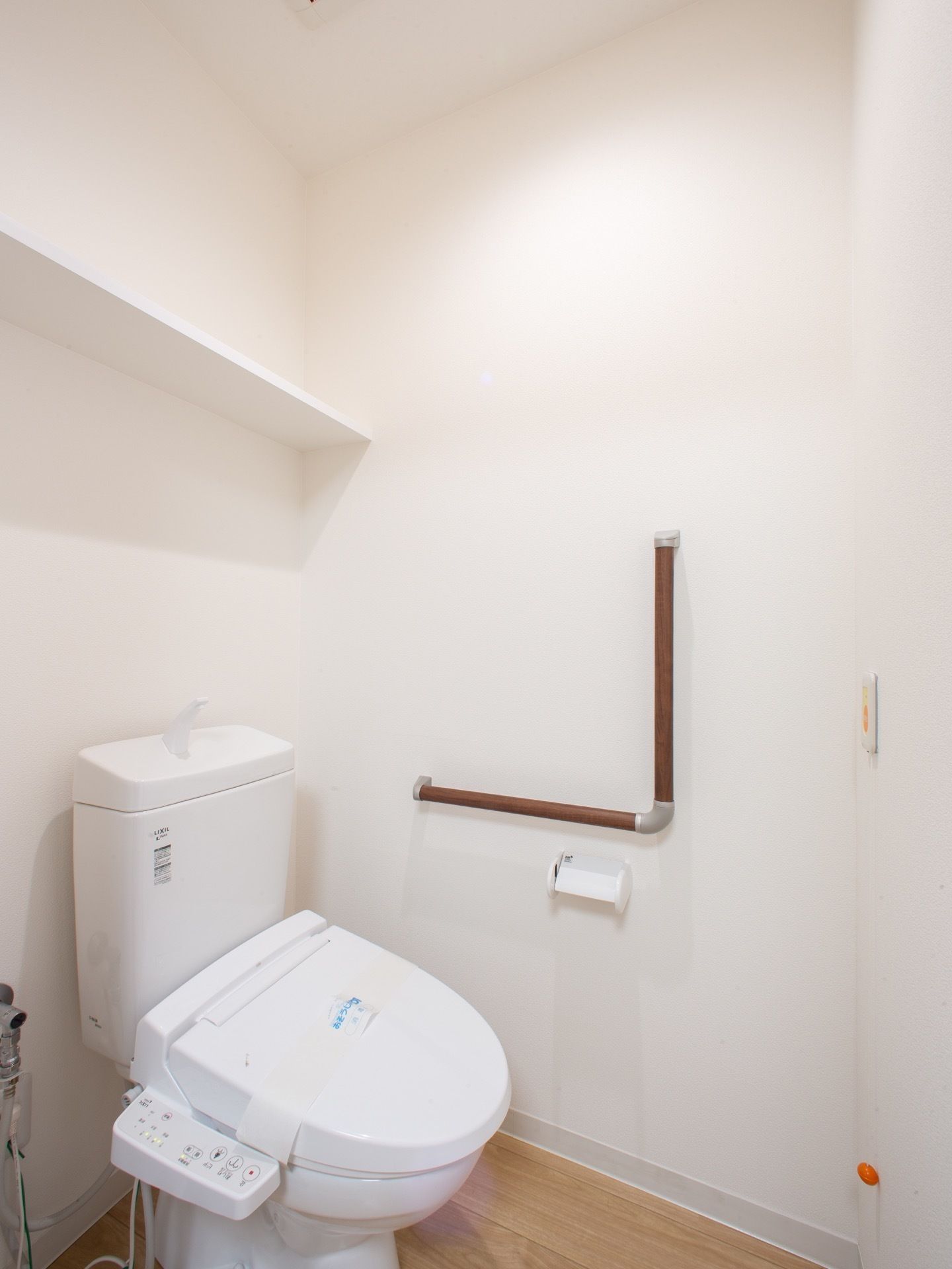 温水洗浄便座が設置されたトイレである。新品を表す紙が蓋に取り付けられている。壁にはL字型に手すりがある。